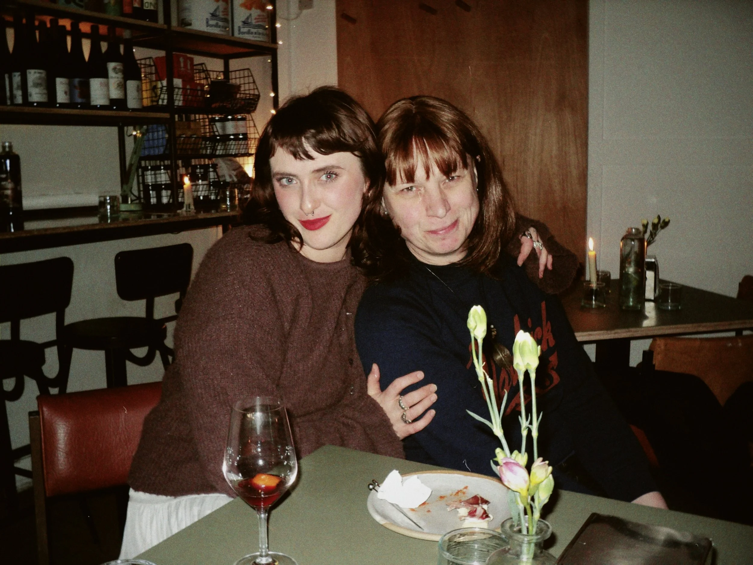 Two women sitting at a dining table in a restaurant smiling for the camera. One has short dark hair and the other has shoulder-length brown hair. There are flowers in a vase, a glass of red wine, and some food on the table.