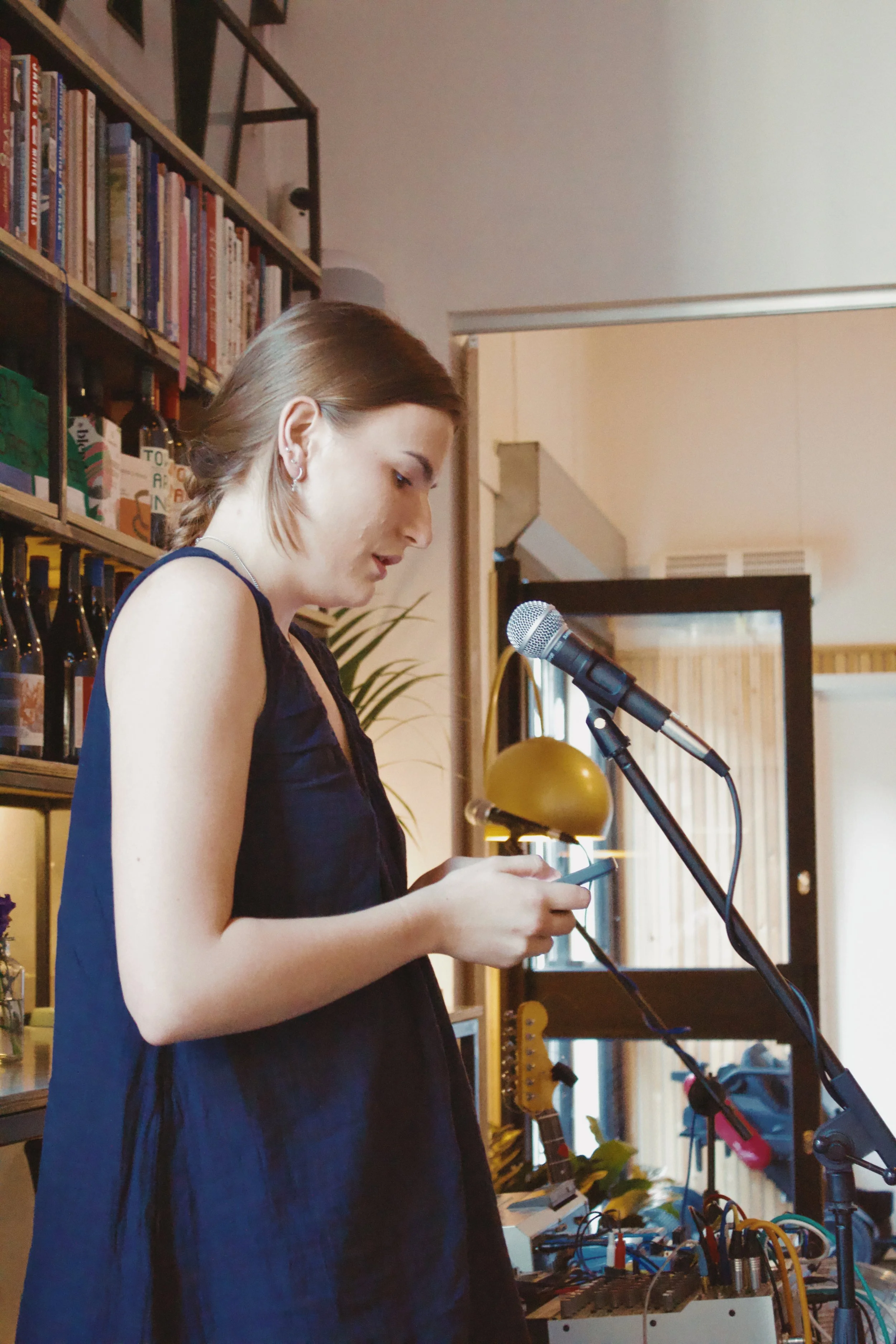 A young woman with shoulder-length brown hair, wearing earrings and a dark blue dress, is standing in front of a microphone while looking at her smartphone. She is in a room with bookshelves, musical instruments, cables, and electronic equipment visi