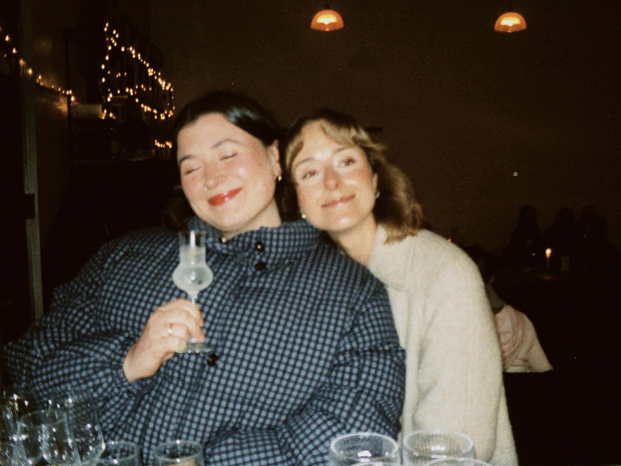 Two women smiling at a gathering, holding drinks, with string lights and lamps in the background.