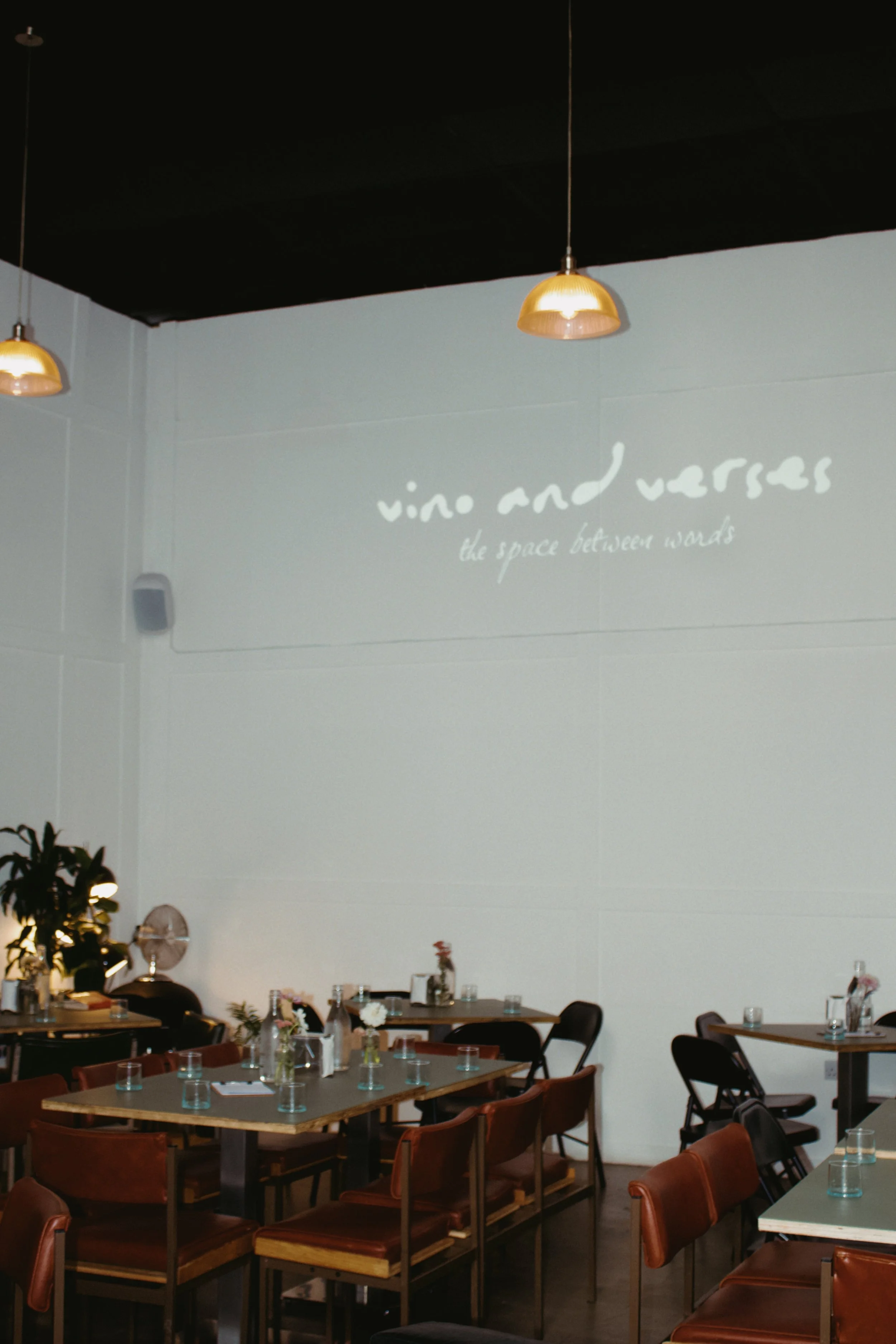 Interior of a restaurant with multiple tables set with glasses and small flowers in vases, with a large white wall displaying the phrase 'vino and verbs' and 'the space between words' in cursive. Hanging lights provide a warm glow.