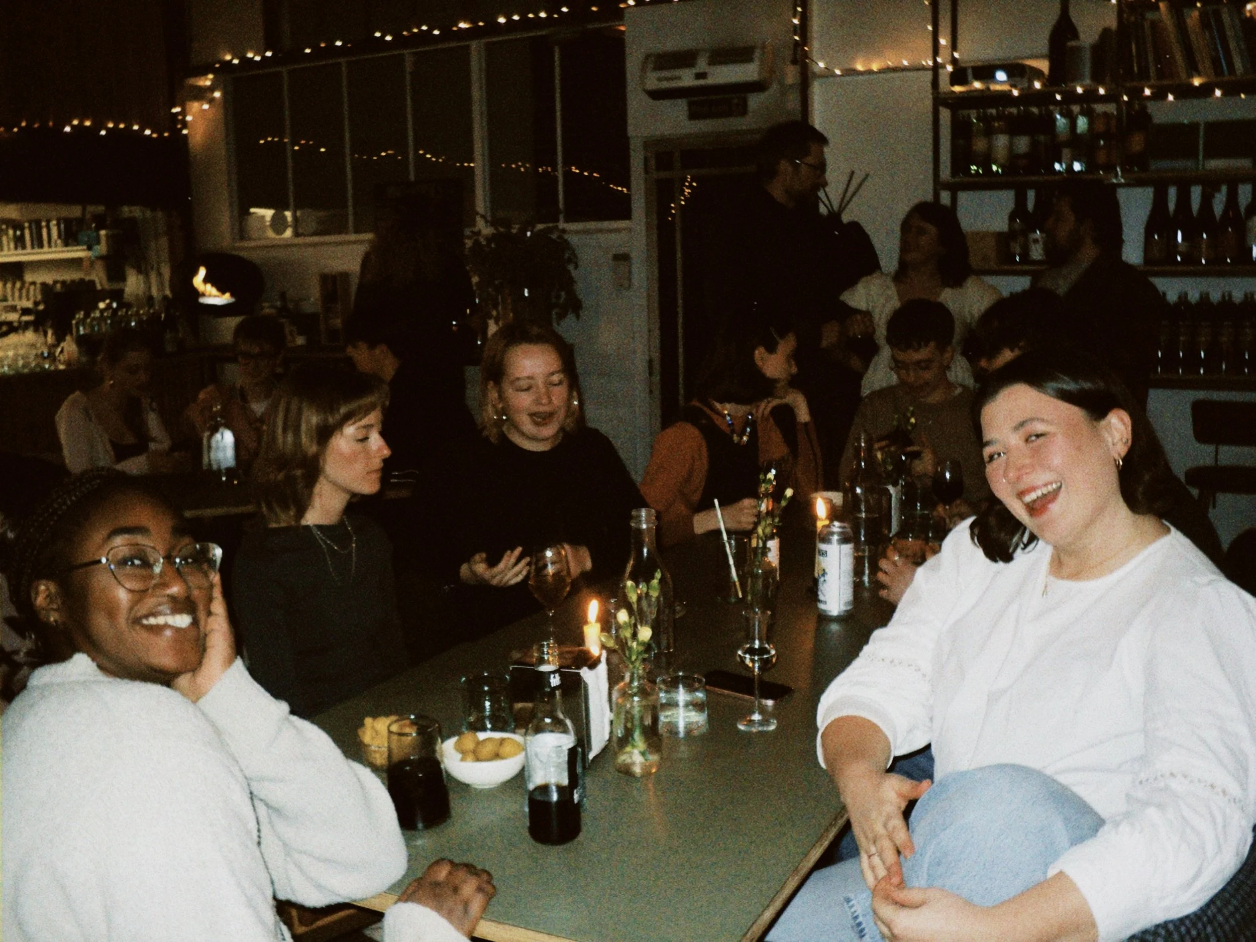 People sitting around a dining table in a dimly lit restaurant celebrating, with some individuals smiling, holding drinks, and engaging in conversation.