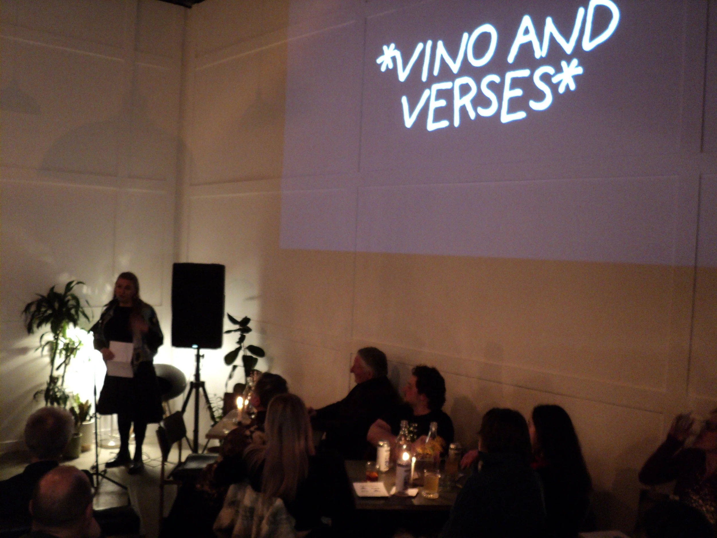 A woman is standing on a small stage or platform, speaking into a microphone, in a dimly lit room decorated with plants. An audience is seated at tables with candles or small lights, watching her. A large illuminated sign with the text '#VINO AND VER