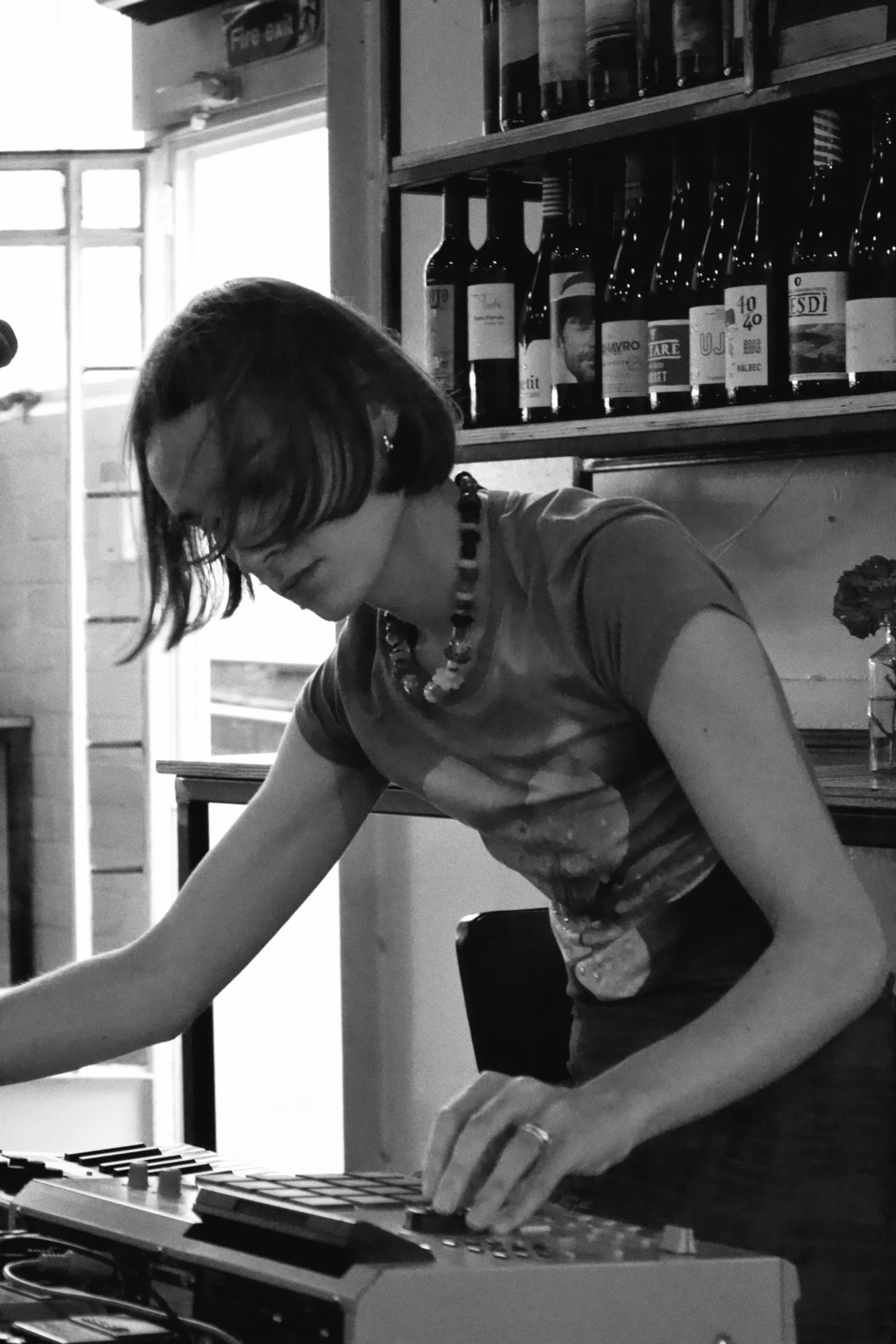 A person with short hair and glasses working with an electronic device on a table in a room with a shelf of bottles in the background.