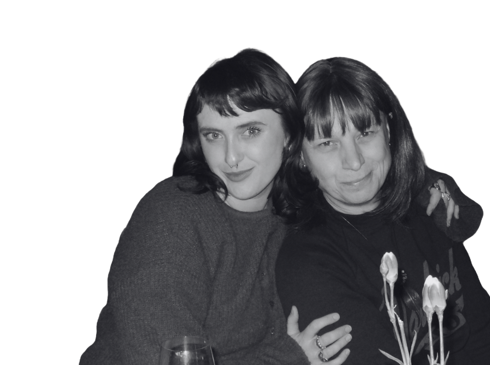 Two women with dark hair and bangs, smiling, sitting close together with arms around each other, in black and white
