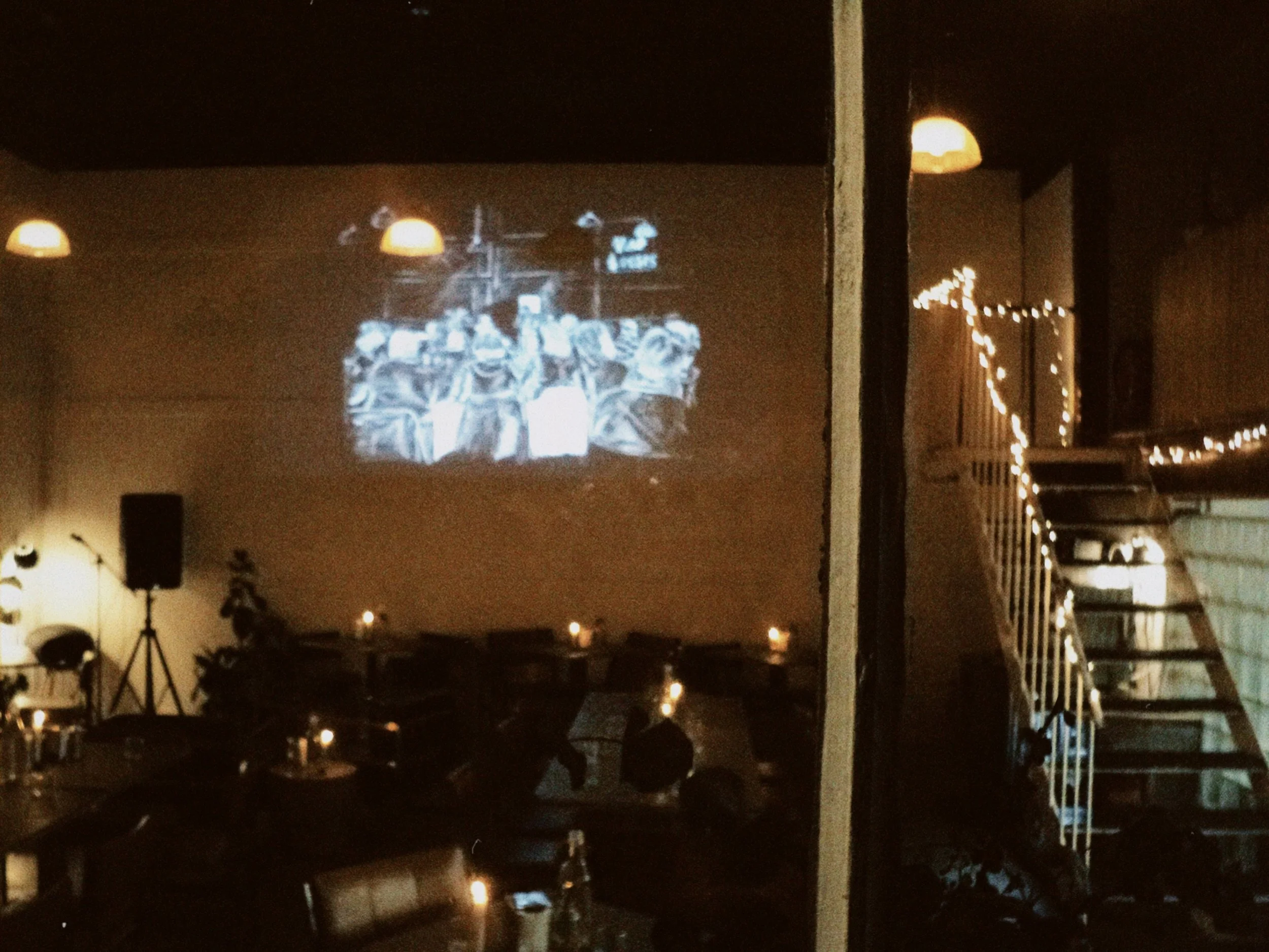A living room with a wall-mounted TV. The TV is displaying a black-and-white photo of a group of people. The room has dim lighting with a few ceiling lights and small string lights along a balcony railing. There are several chairs and tables in the r