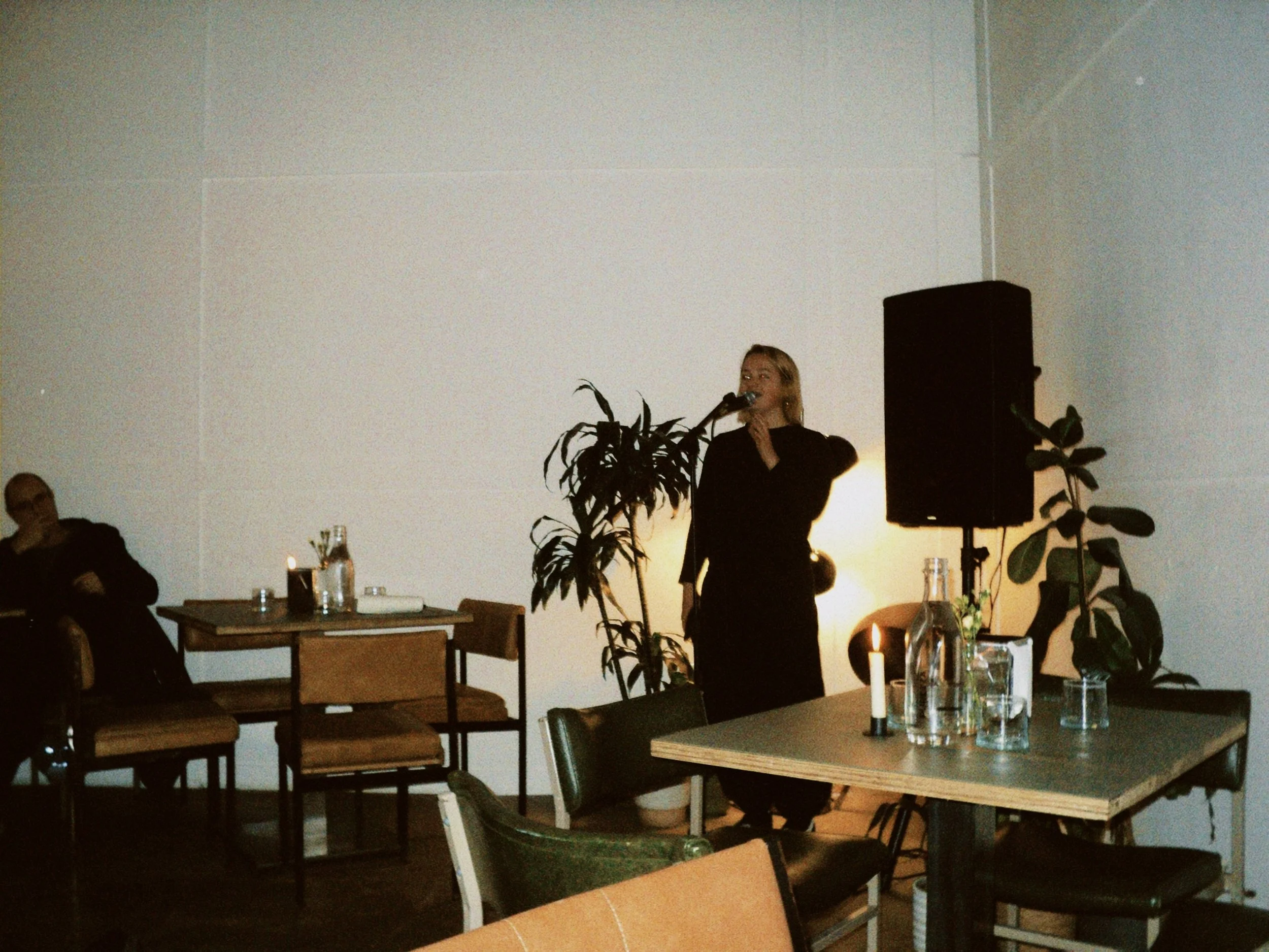A woman speaks into a microphone while standing in a dimly lit room decorated with plants and candles, with some people seated at tables.