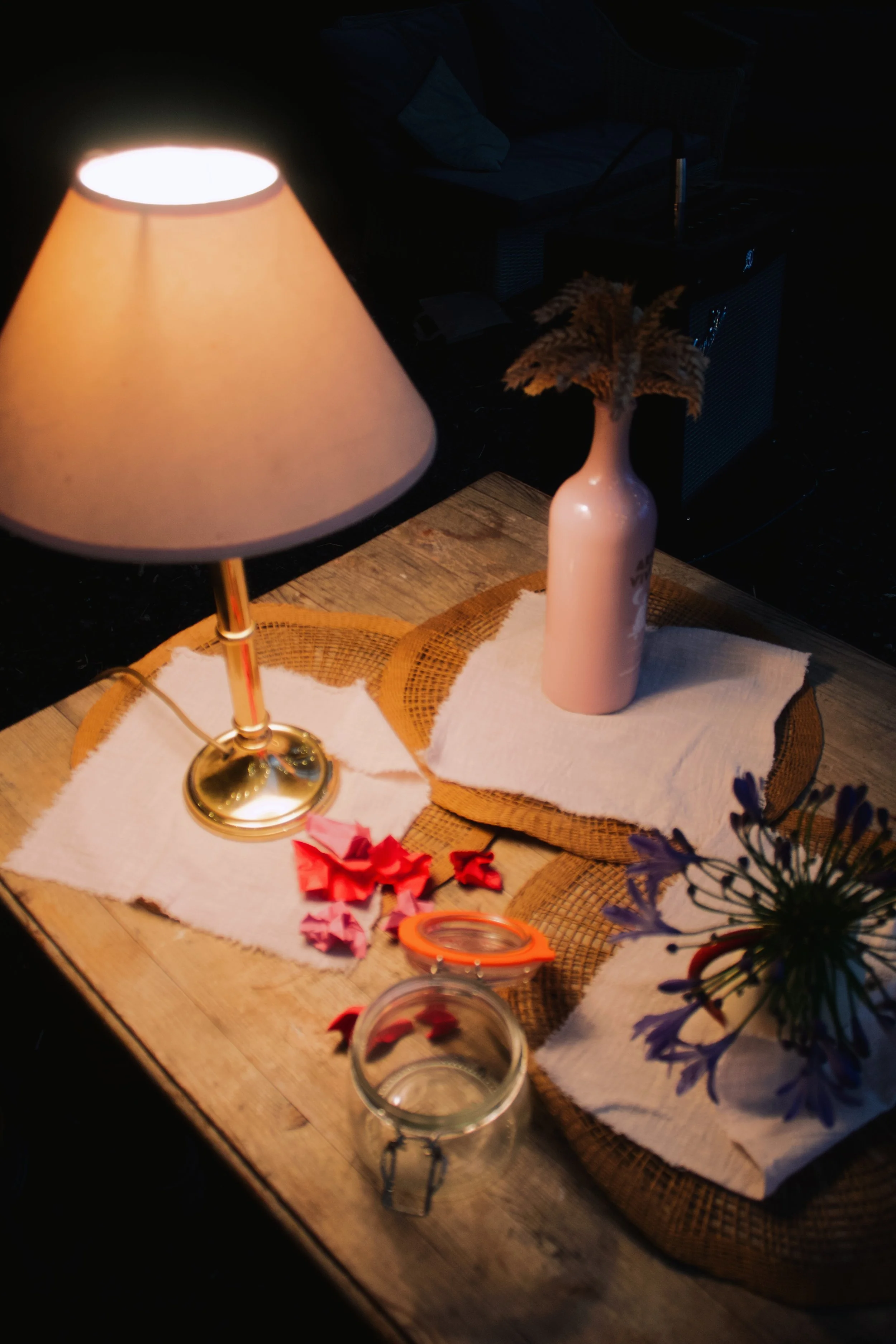 Close-up view of a wooden table with a white cloth runner, a lamp with a beige shade turned on, a pink vase with dried flowers, a glass jar with a lid, and some red and pink paper decorations. Blurred background with a sofa and other objects.