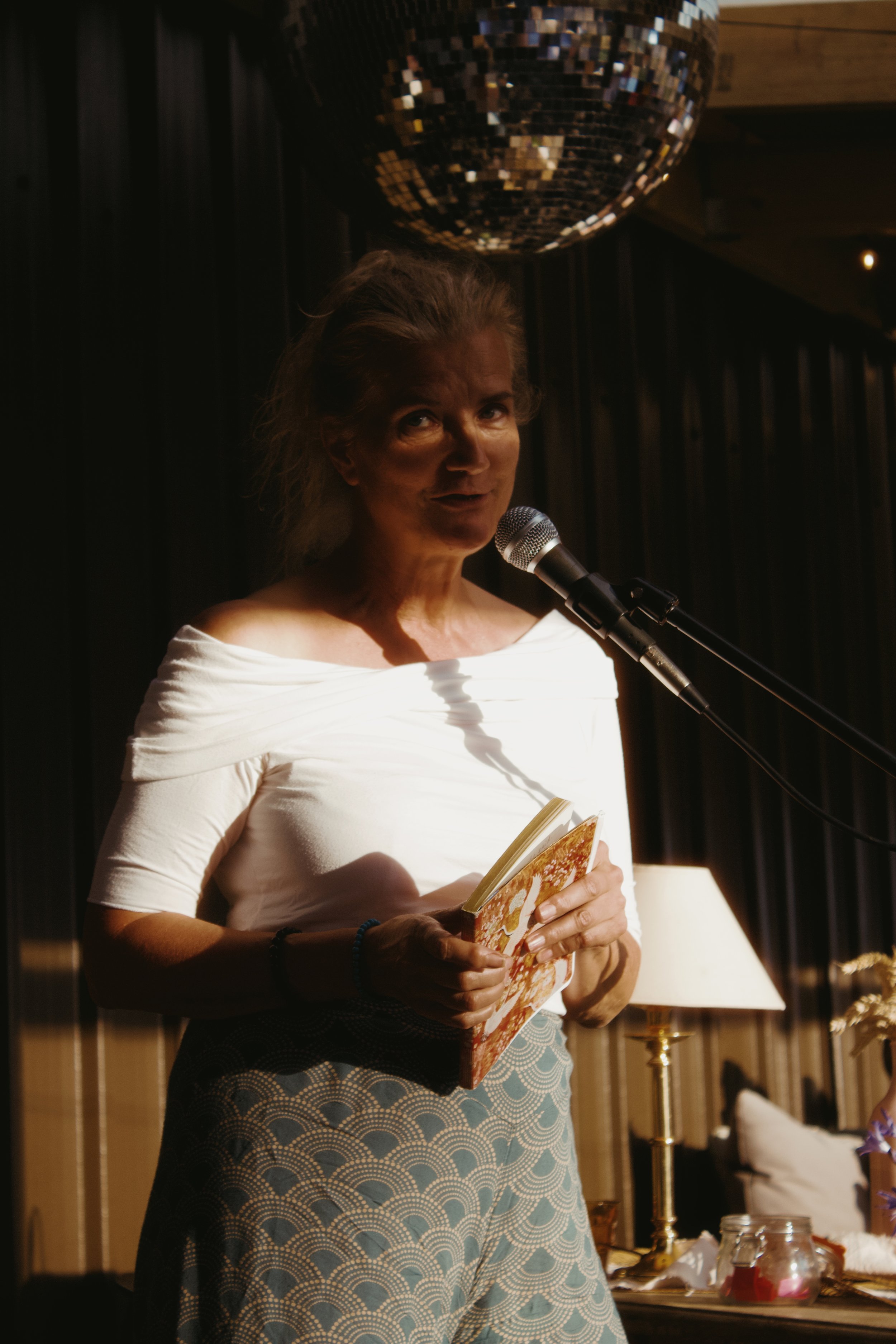 A woman with gray hair wearing a white off-the-shoulder top and patterned skirt, standing near a microphone, holding a notebook, in a dimly lit room with dark curtains, a lamp, and decorative jars on a table.
