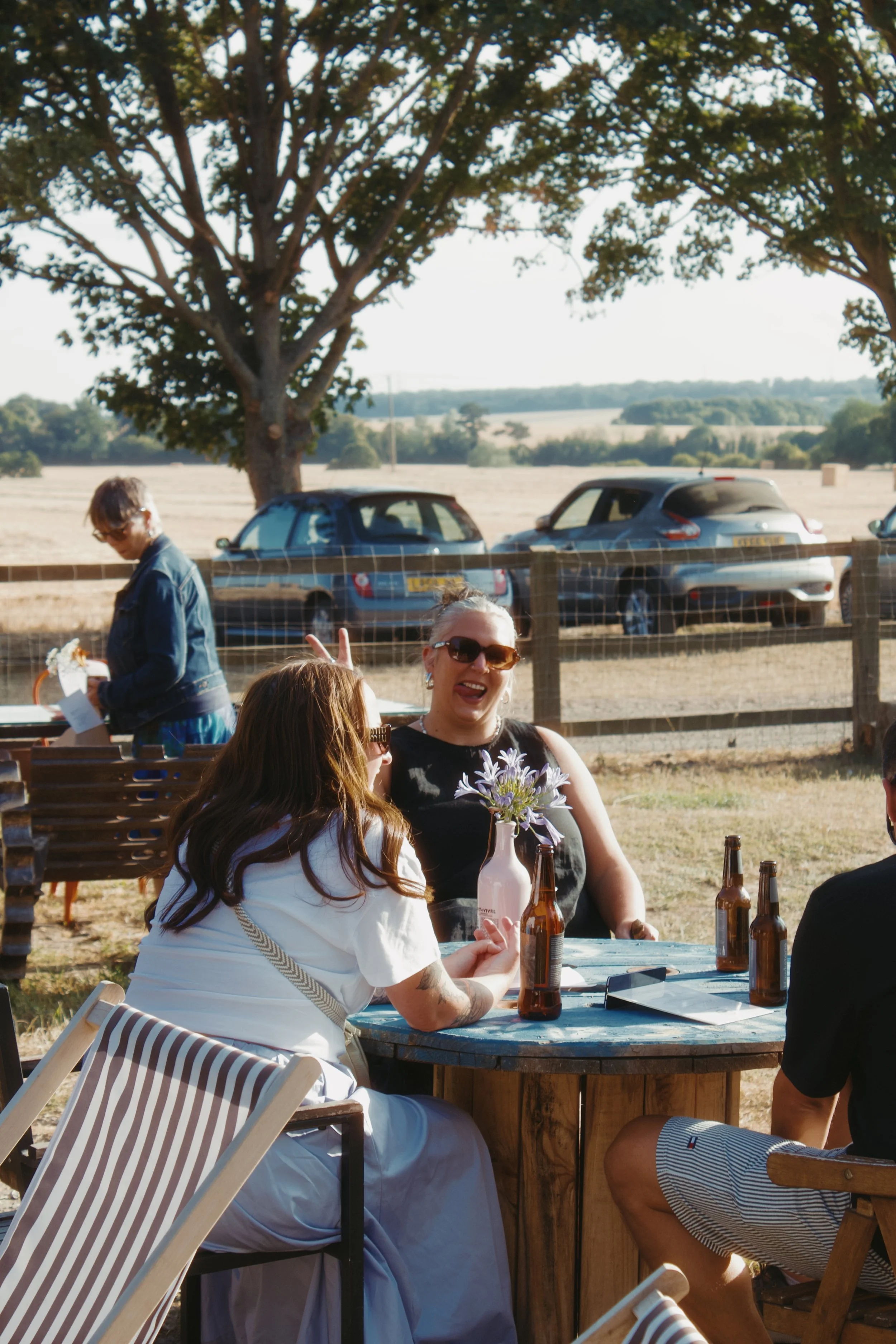 People sitting at a wooden table outdoors, enjoying drinks and conversation under a large tree on a sunny day.