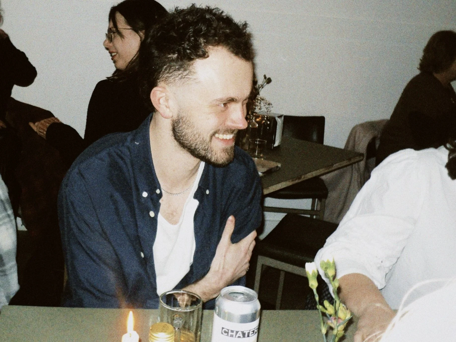 A man with curly hair and a beard smiling and crossing his arms, sitting at a table during a gathering. There are people in the background and drinks on the table.