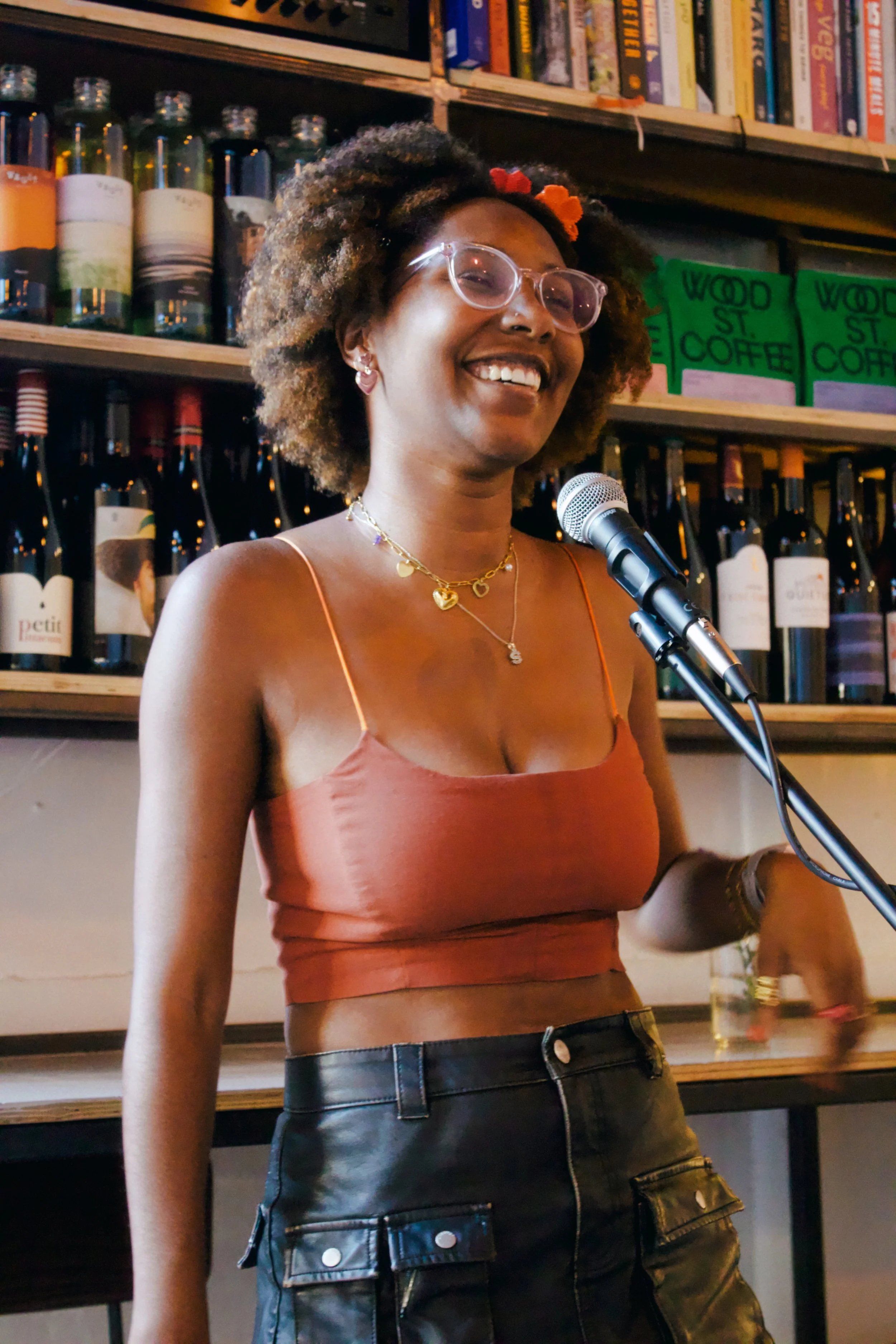 A woman smiling and speaking into a microphone indoors, with bookshelves and liquor bottles in the background.