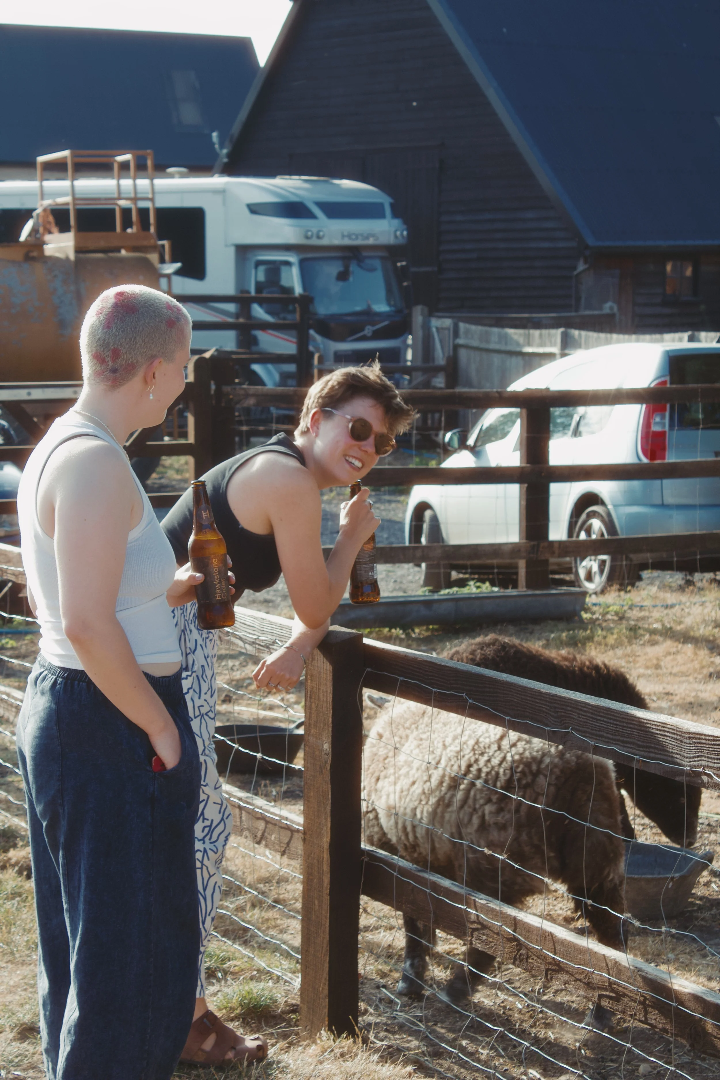 Two women standing by a wooden fence at a farm, watching a large brown sheep. One woman has short pink hair and a white sleeveless top, the other has short brown hair, sunglasses, and a black sleeveless shirt. They are holding beer bottles and smilin