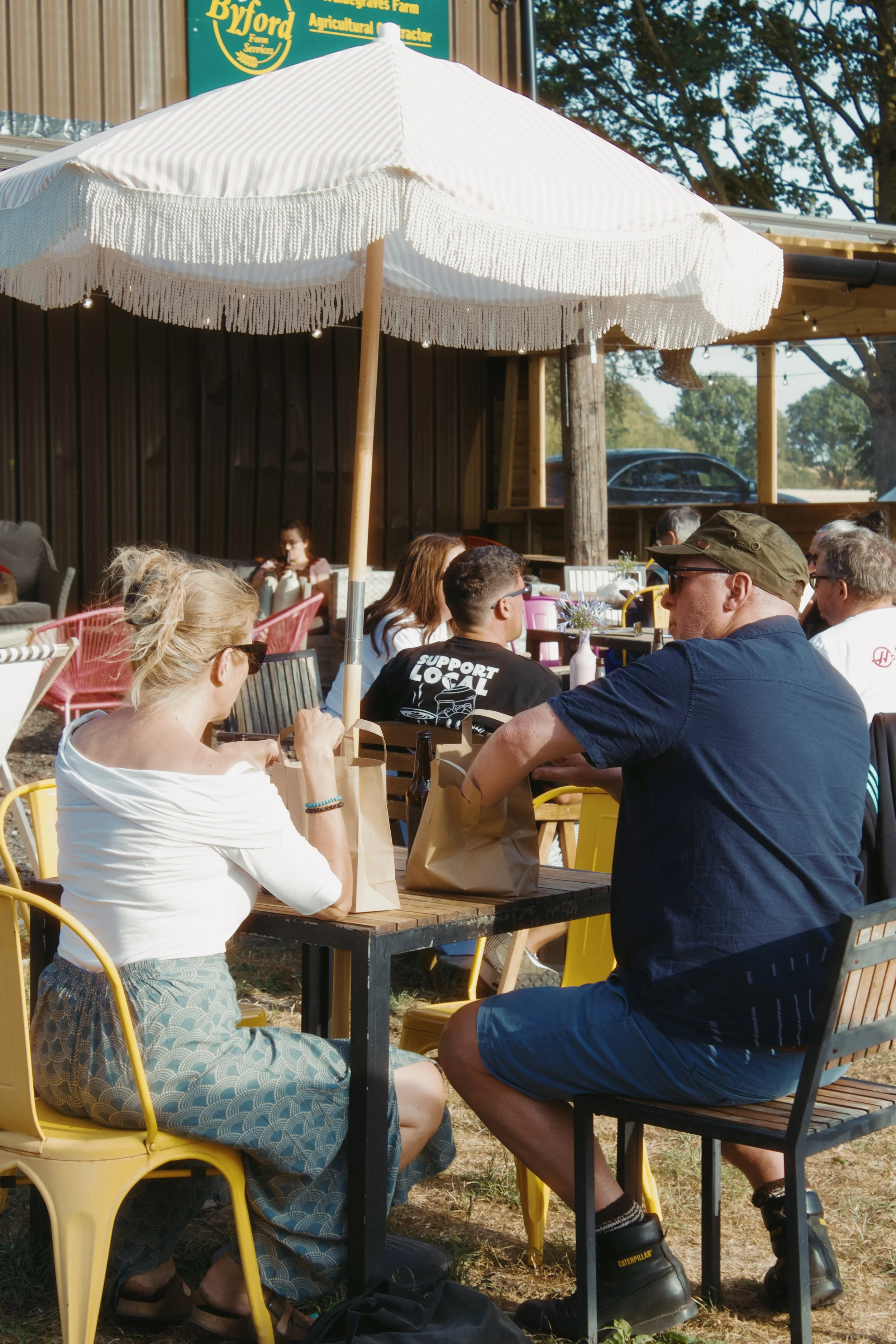 People sitting outdoors under a large white patio umbrella, socializing at tables with brown paper bags, in front of a wooden building with a sign that reads 'Byford Farms, Agricultural'.