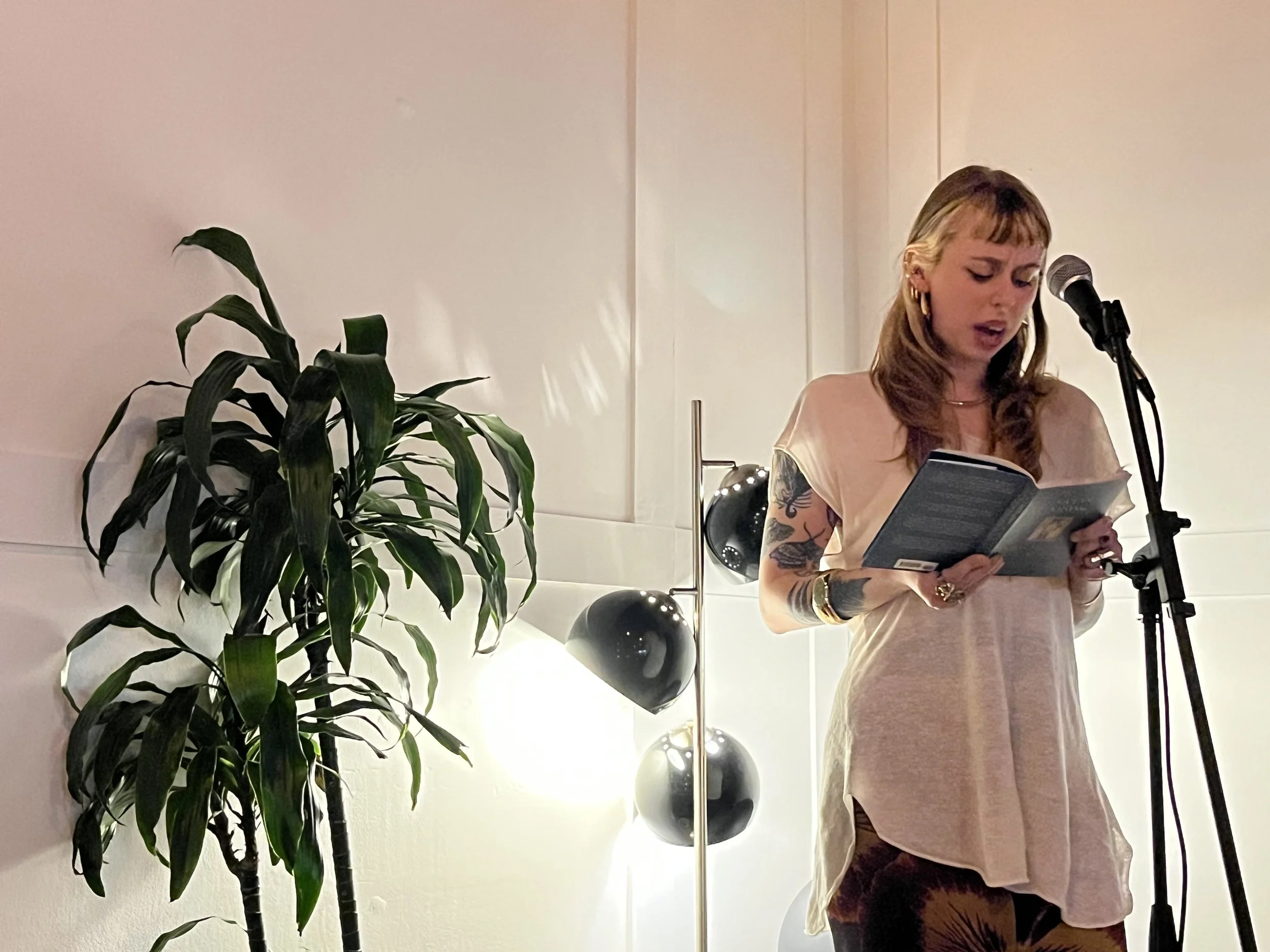 A woman with shoulder-length blonde hair and tattoos on her arms, wearing a light-colored dress, is reading from a book and singing into a microphone, positioned beside a tall green plant and a modern lamp with multiple round bulbs.