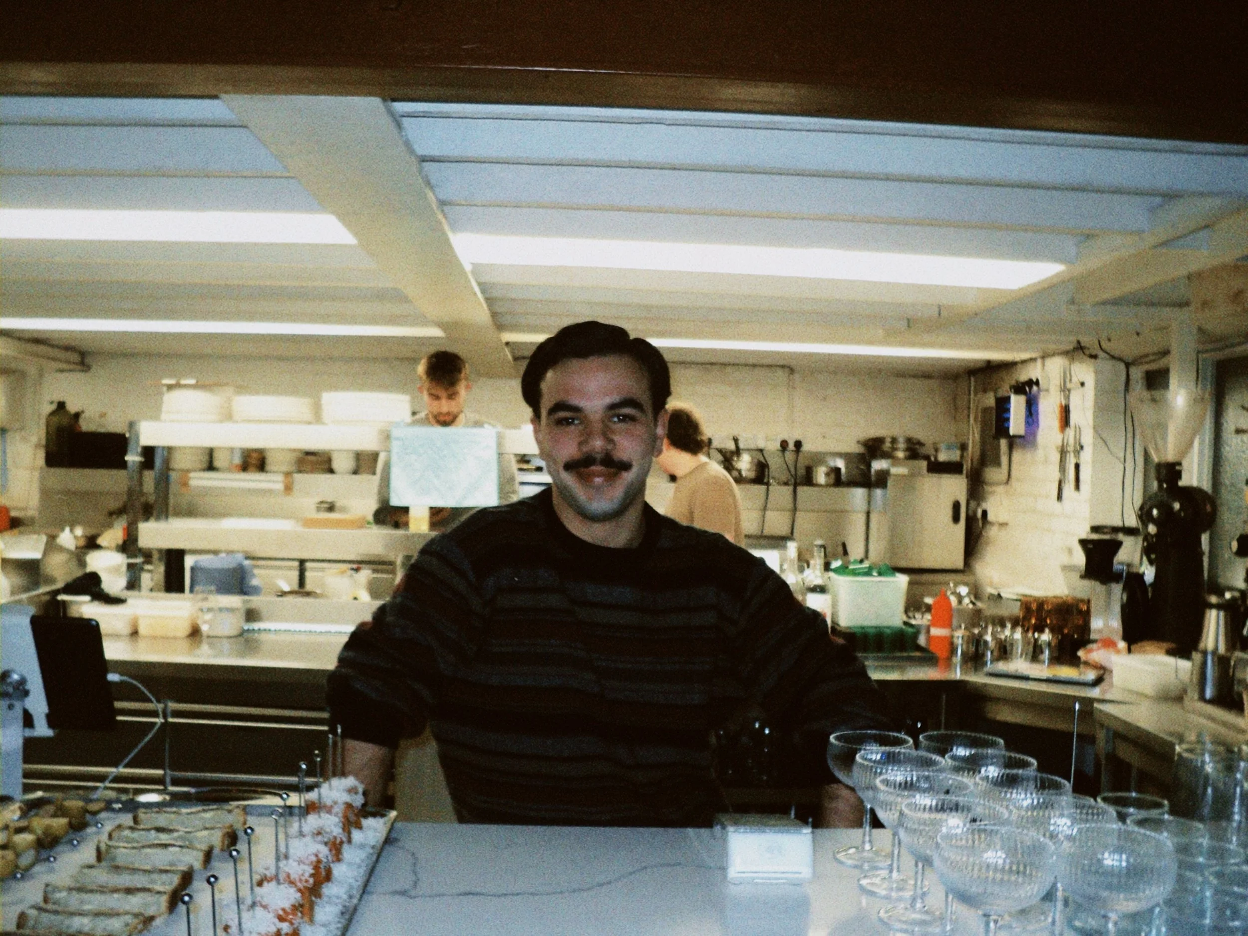 A man with dark hair and a mustache sitting at a table in a busy industrial kitchen, smiling at the camera. There are other people working in the background, along with various kitchen equipment and supplies, including trays of appetizers, glass cups