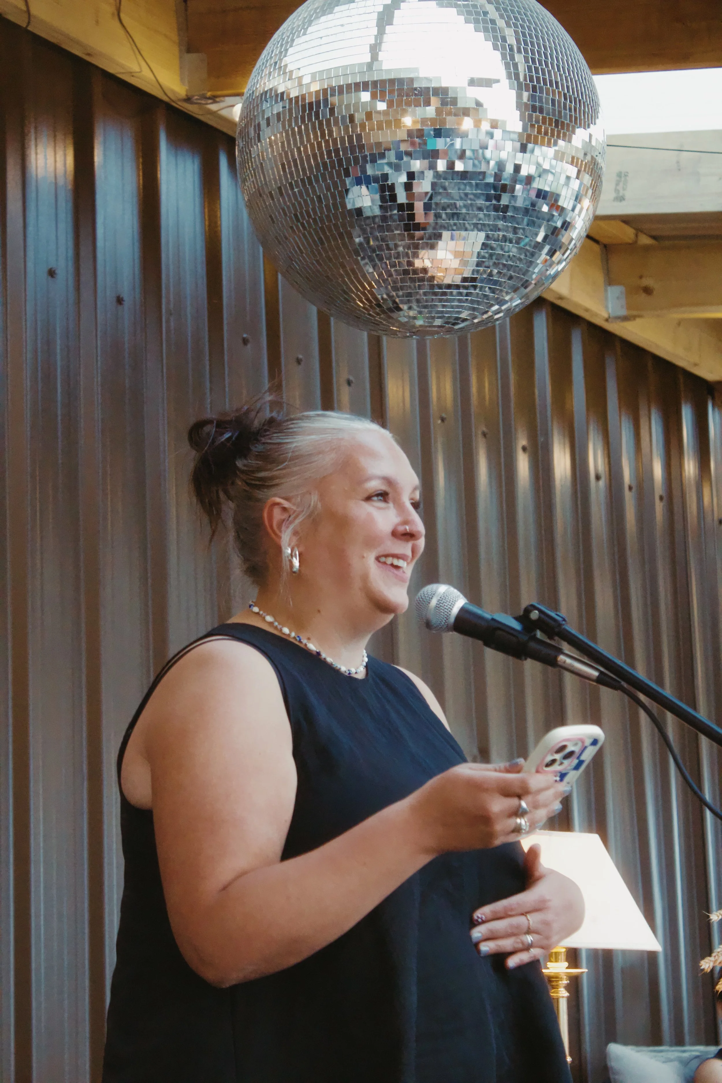A woman with blonde hair tied back in a bun, wearing a black sleeveless dress, earrings, a necklace, and several rings, standing indoors with wood paneling background. She is smiling and holding a smartphone while speaking into a microphone in front 