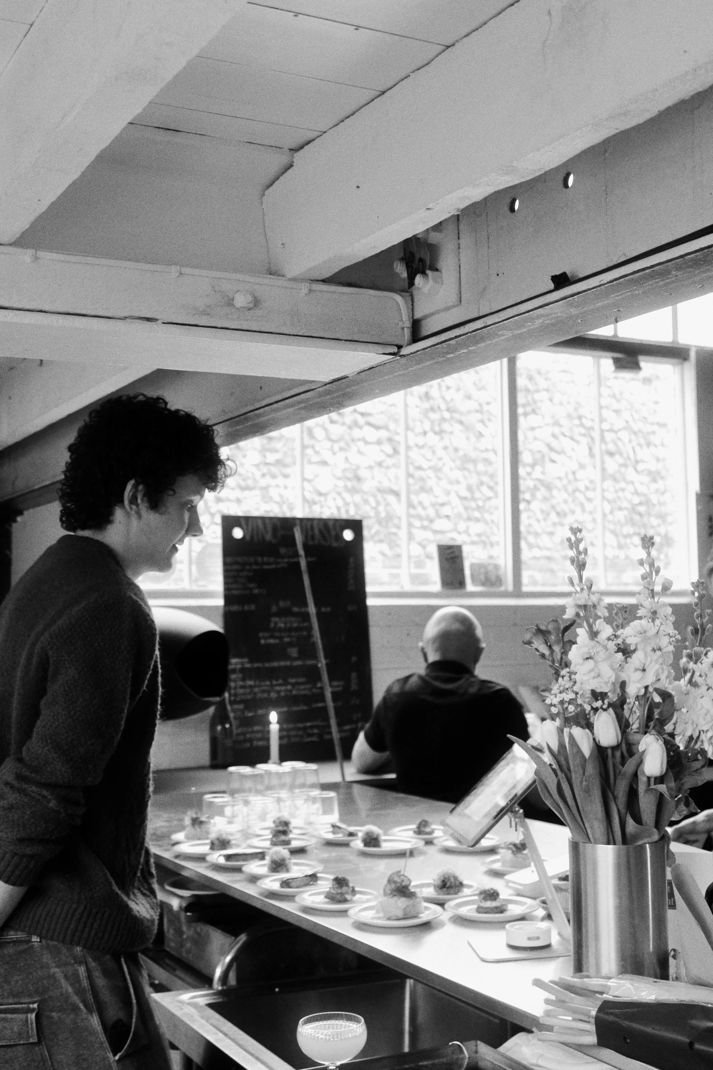 A woman standing at a table with small plates of food, in a room with large windows and flowers in a vase, with other people in the background.