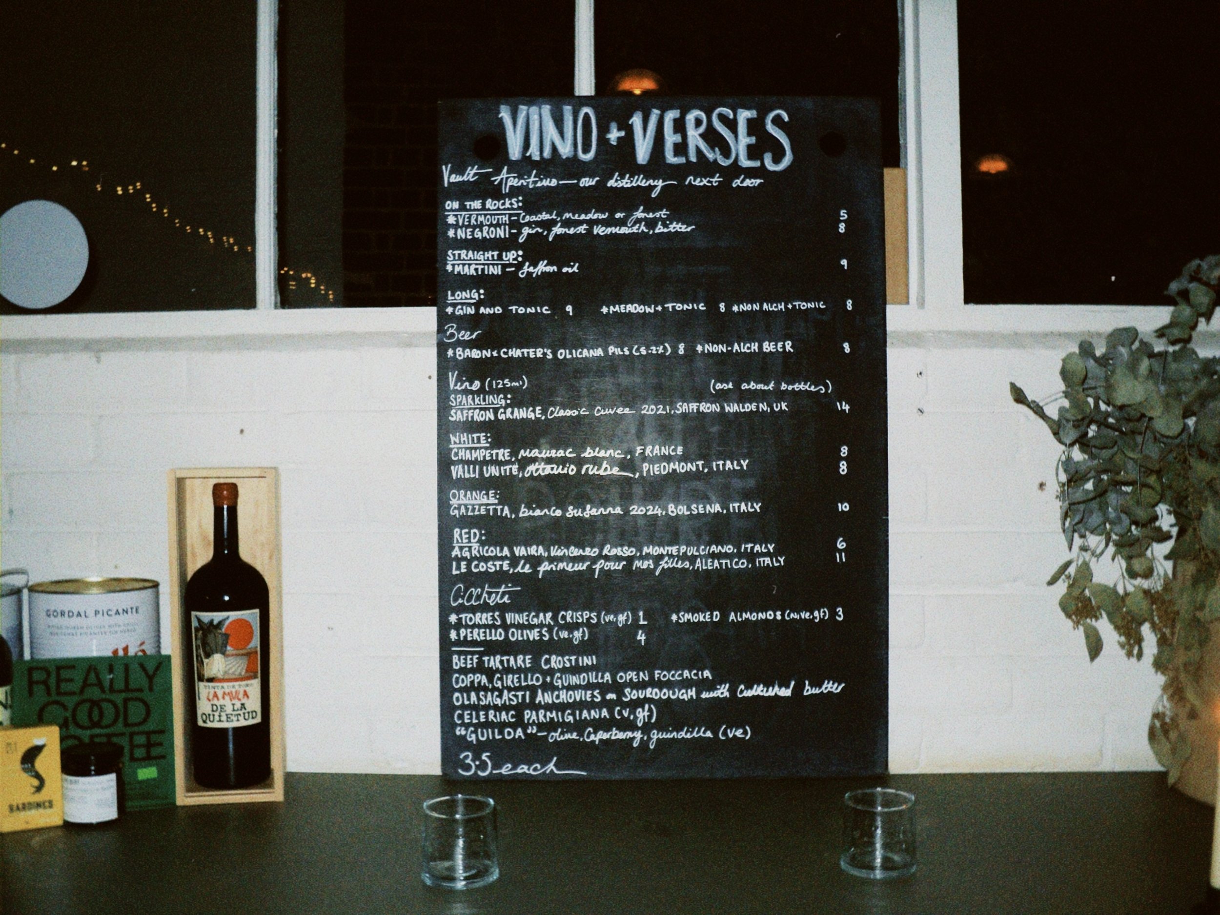 Black chalkboard menu titled 'Vino + Verses' listing various wines, beers, and snacks, with glasses in front and bottles and signage nearby.