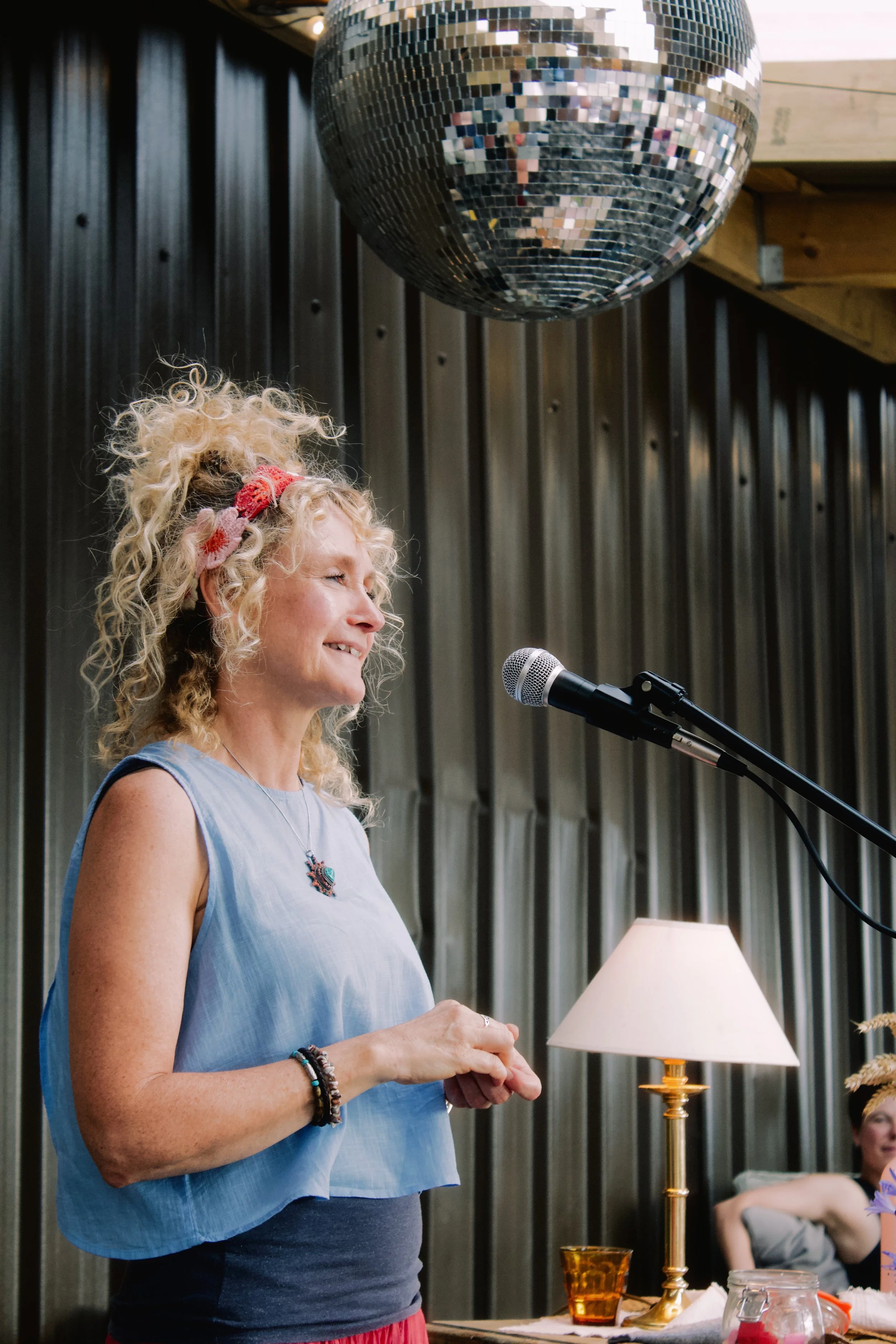 A woman with curly blonde hair wearing a light blue top and jewelry, smiling while standing next to a microphone. A disco ball hangs above her. In the background, a dark paneled wall and a person sitting on a gray couch are visible, with a table lamp