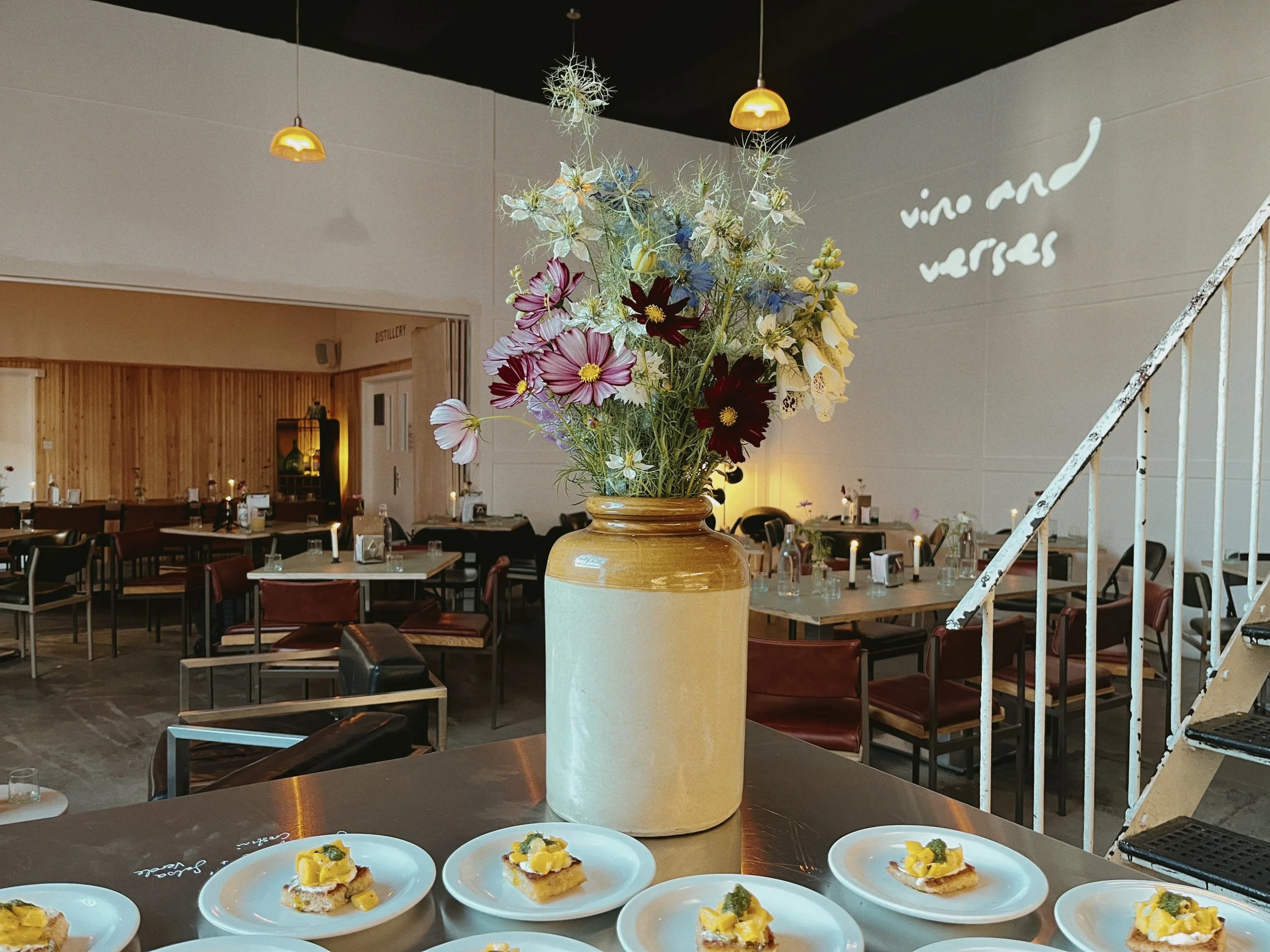A large ceramic vase with a bouquet of assorted flowers including cosmos, clematis, and other wildflowers. The vase is placed on a table in a decorated restaurant setting with tables and chairs, pendant lighting, and wall decor in the background.