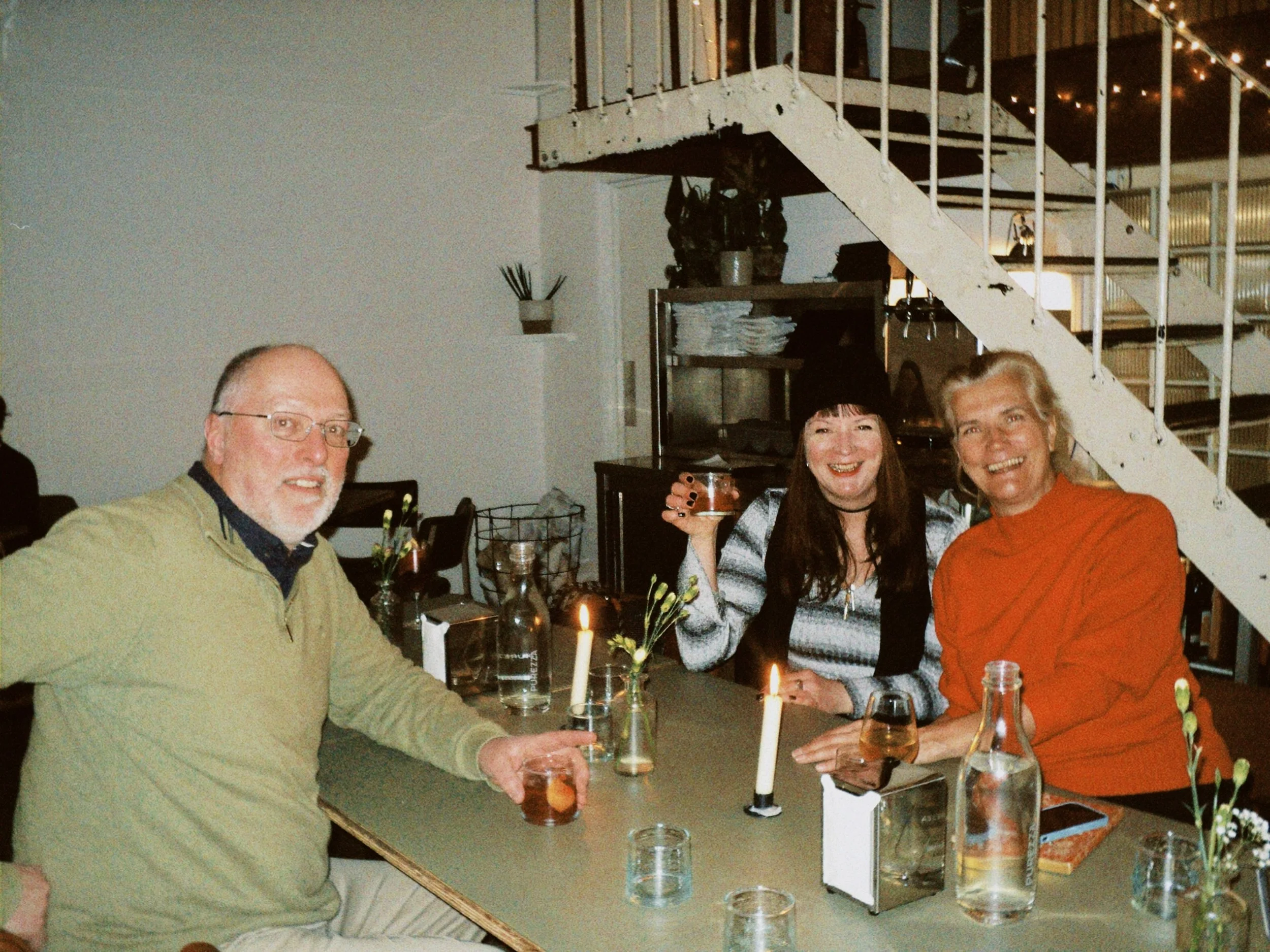 Three people sitting at a dining table with candles and drinks, smiling at a gathering in an indoor setting, with a staircase visible in the background.