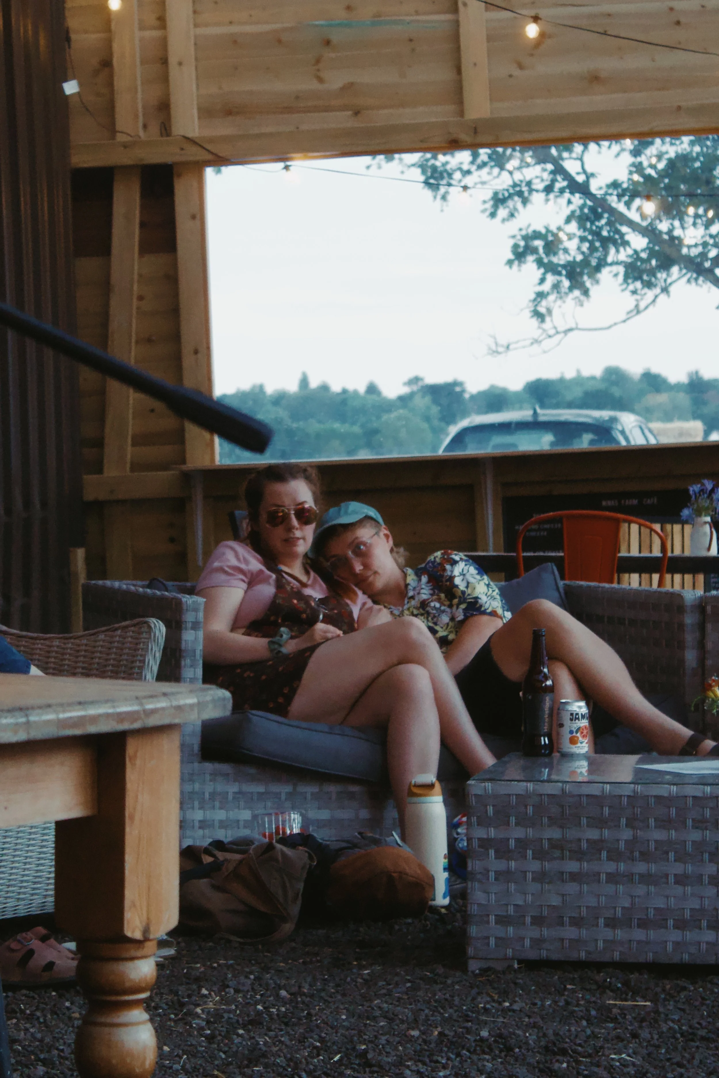 Two women sitting on a gray wicker outdoor sofa in a wooden pavilion, with a person leaning on them. They are in a relaxed setting, with drinks on a table in front and bags on the ground. The background shows trees, a car, and string lights hanging o