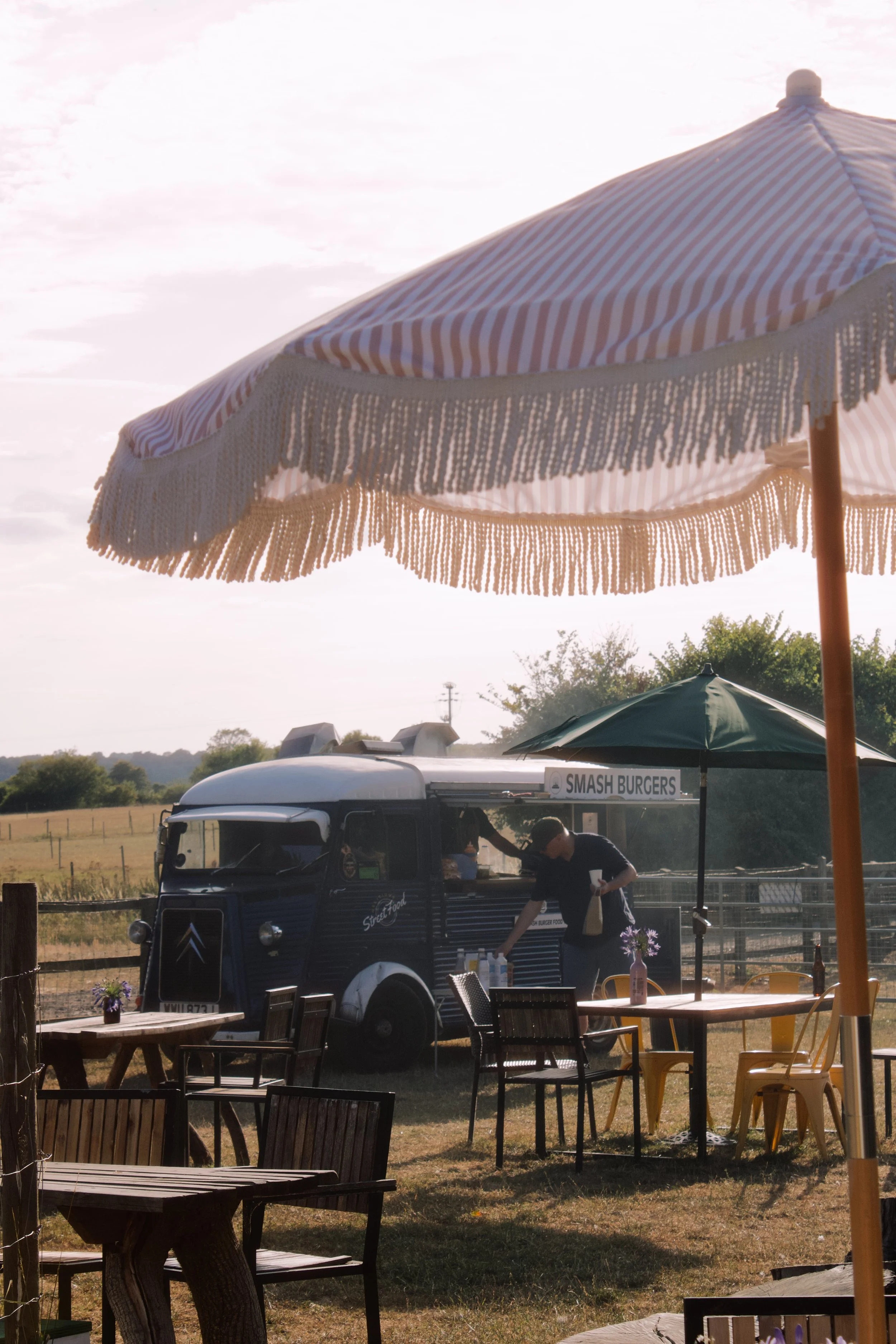 Outdoor setup featuring a striped beach umbrella with fringe, various tables and chairs, a street food cart labeled "SMASH BURGERS," and a person preparing food, with trees and open field in the background under a partly cloudy sky.