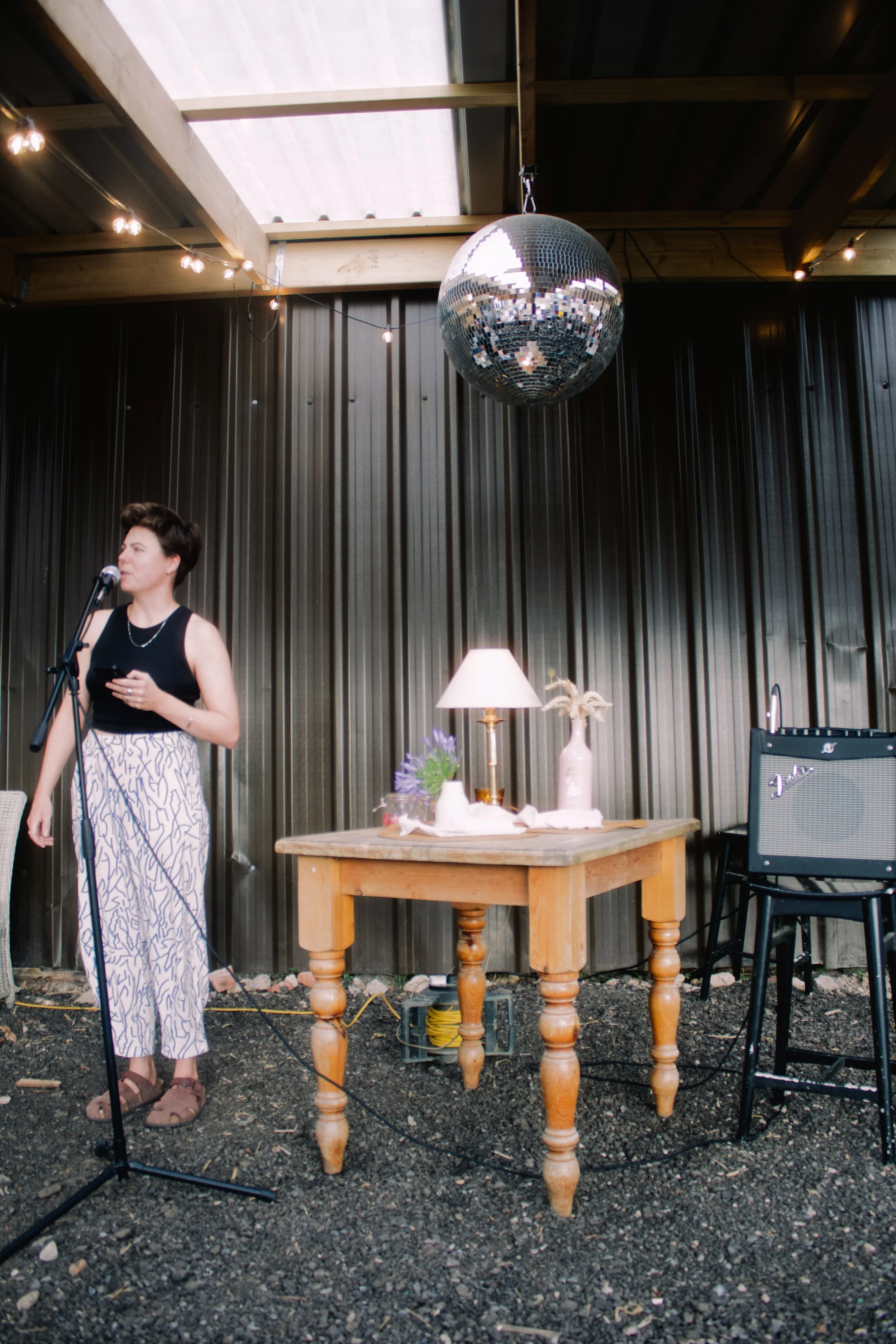 Indoor space with a person standing at a microphone, holding a phone, wearing a black top and patterned pants, in front of a dark wall, with a large disco ball hanging from the ceiling and string lights, a wooden table with decorative items, a lamp, 