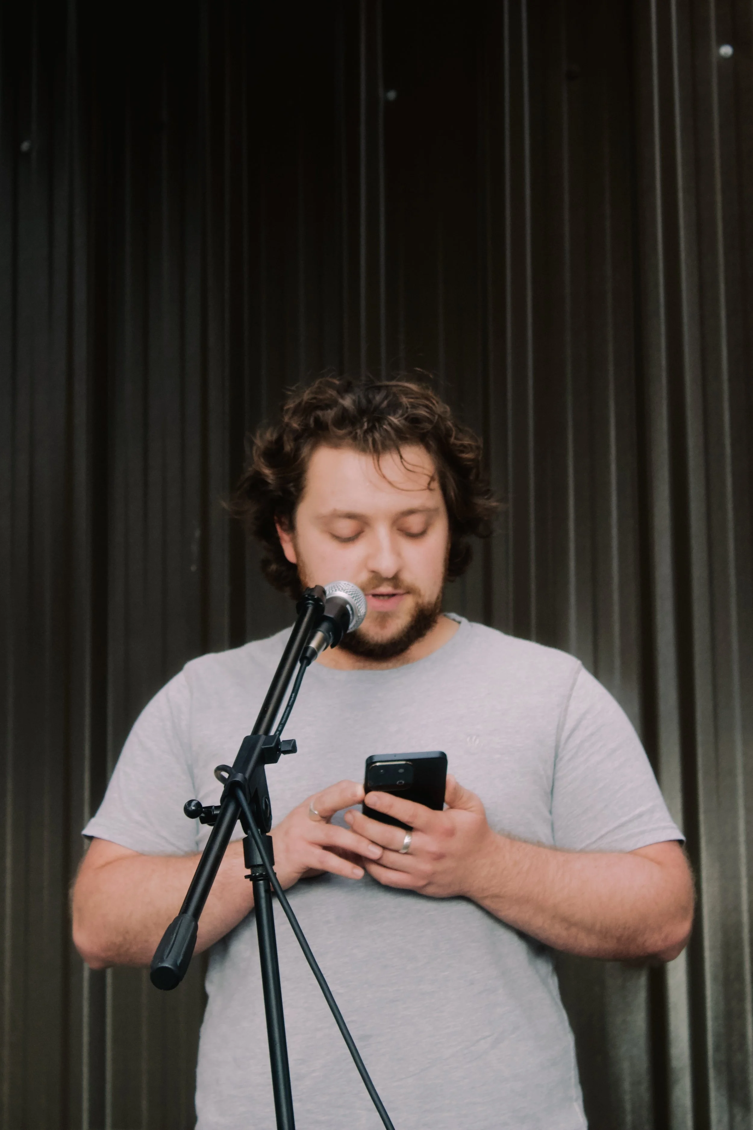 A young man with curly hair and a beard, wearing a light gray t-shirt, standing in front of a black curtain, looking down at his phone, with a microphone on a stand in front of him.