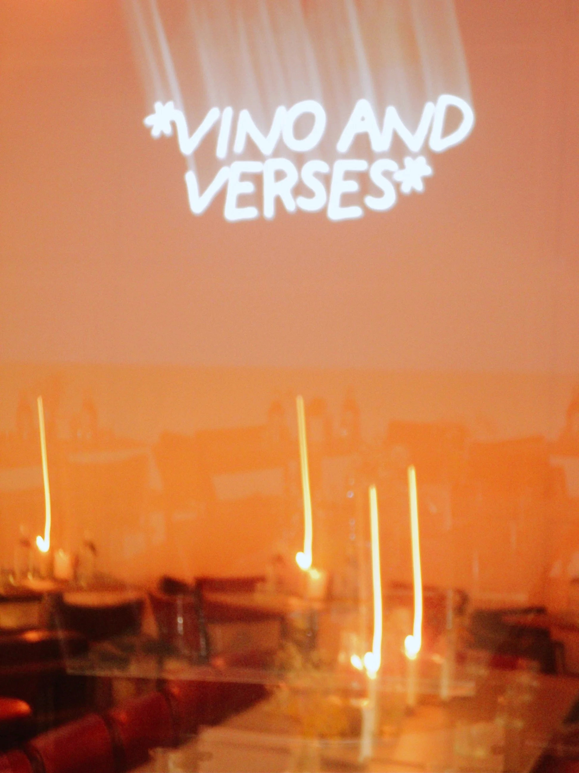 Neon sign with white text on an orange background that says '*VINO AND VERSES*'. In the foreground, there are seating tables and chairs.