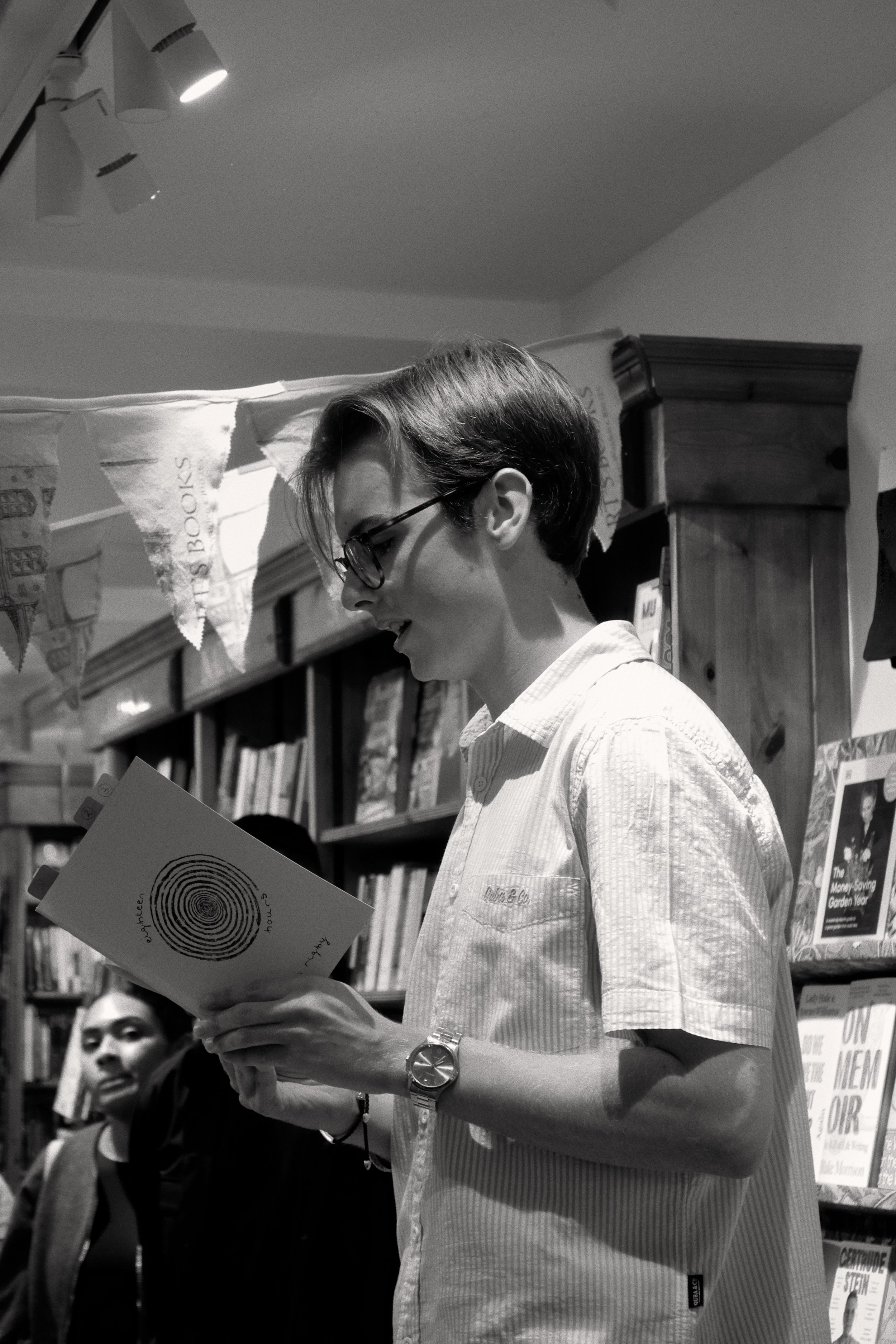 A person with short hair wearing glasses and a checkered shirt, holding a book in a bookstore. There are books on shelves behind them and a woman in the background.