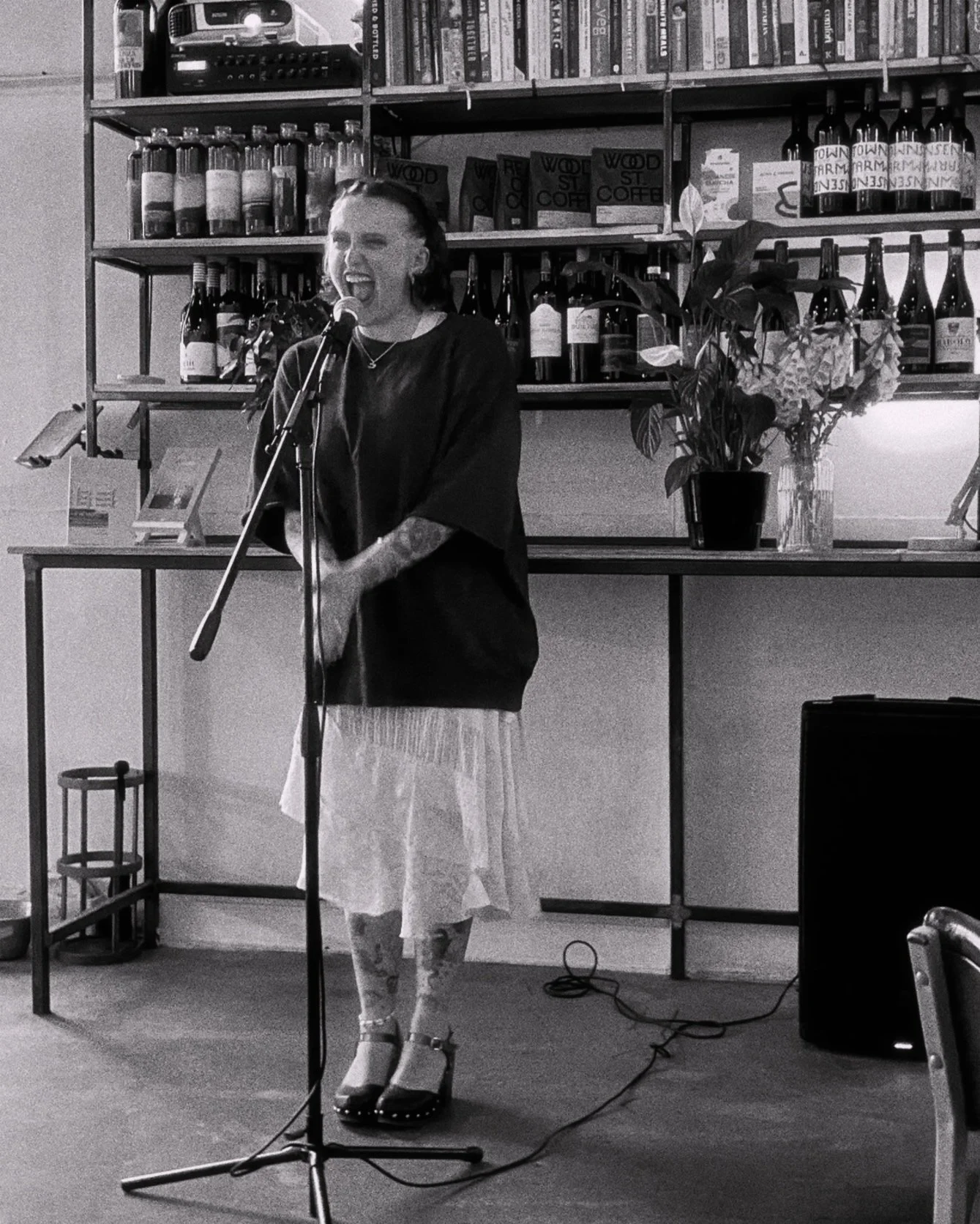A woman singing into a microphone in a cafe, with a shelf of books and bottles behind her.