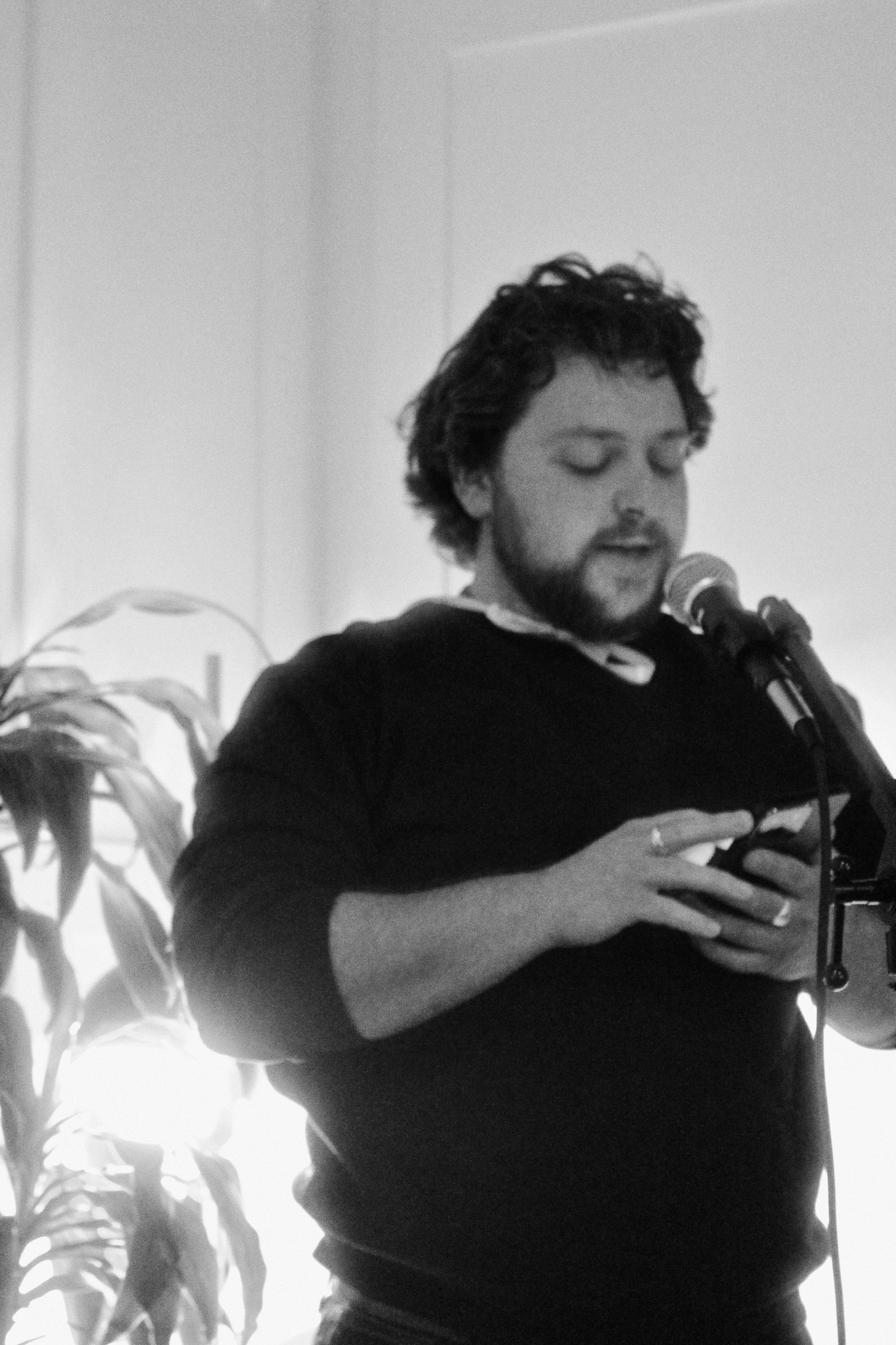 A man with curly hair and a beard sings into a microphone while holding a guitar. He is wearing a dark hoodie, and there are plants in the background.