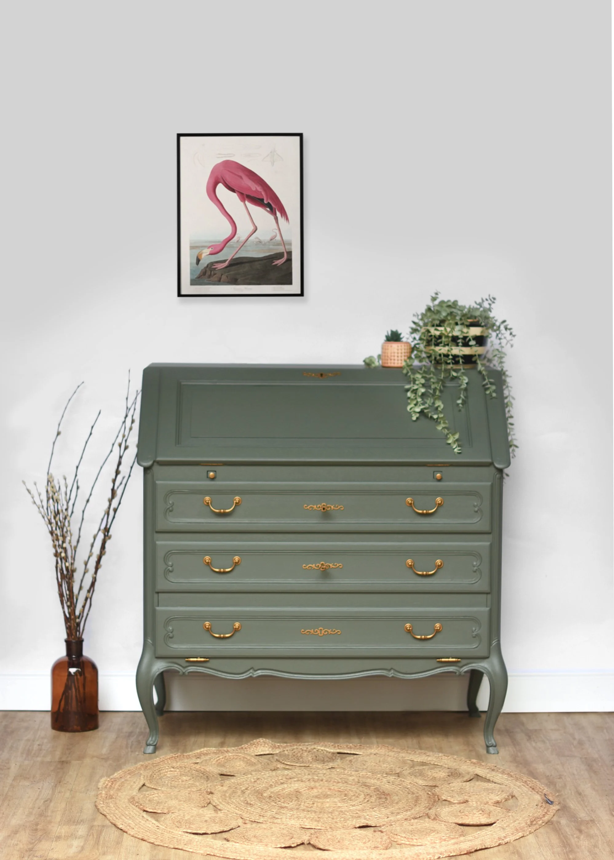 Une commode verte avec des poignées en or, décorée de plantes en pot sur le dessus. À gauche, un vase en verre contenant des branches sèches. Sur le mur, une peinture d'un flamant rose.