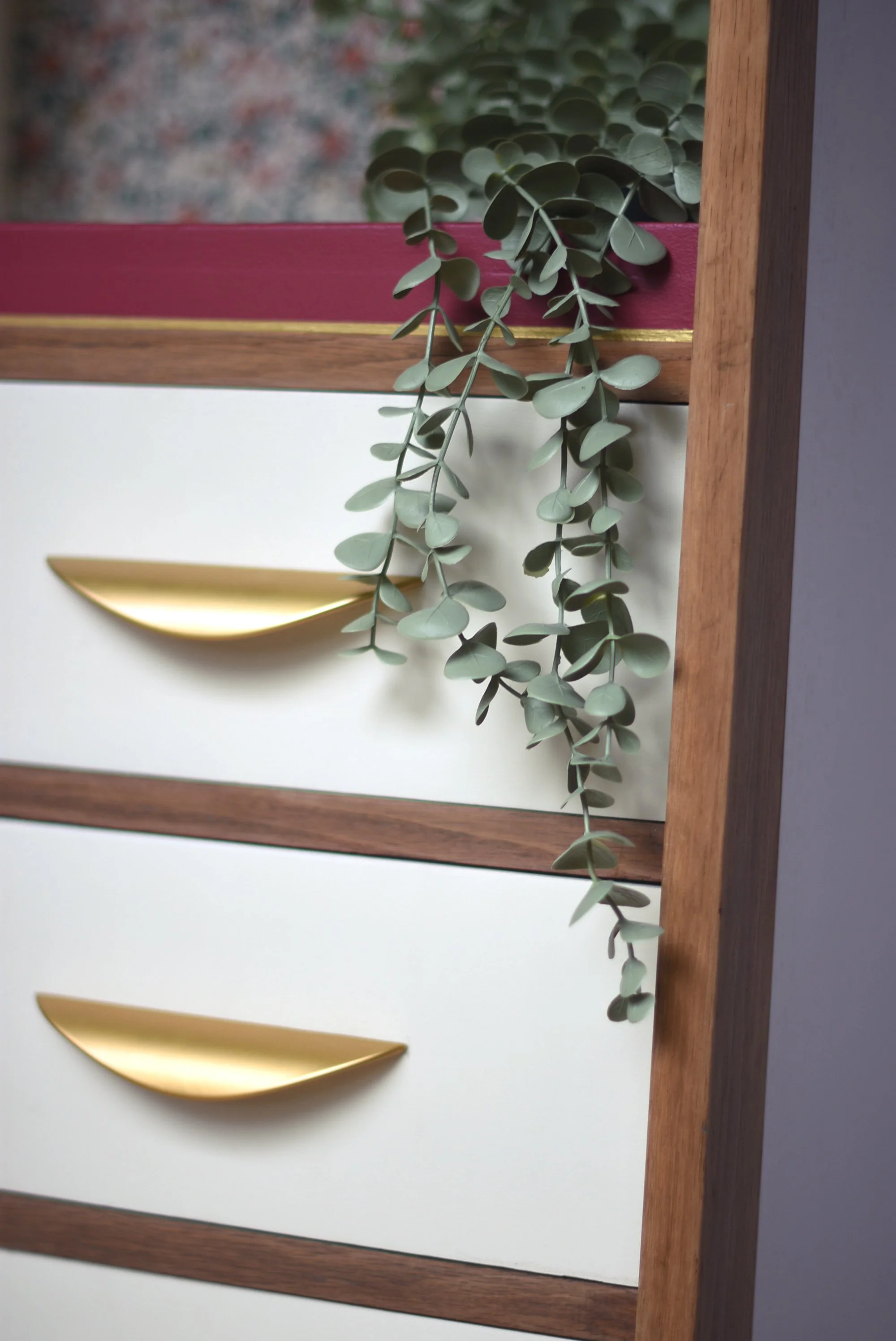Une commode avec trois tiroirs, dont deux blancs avec des poignées dorées, en bois naturel, avec une plante grimpante suspendue à côté.