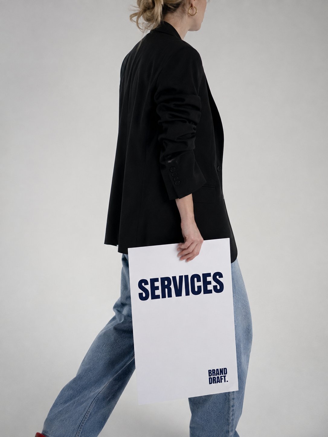 Een persoon in een zwarte jas en blauwe jeans houdt een wit bord met de woorden 'SERVICES' en 'BRAND DRAFT' tegen de zij. Ze staan op een neutrale, lichte achtergrond.