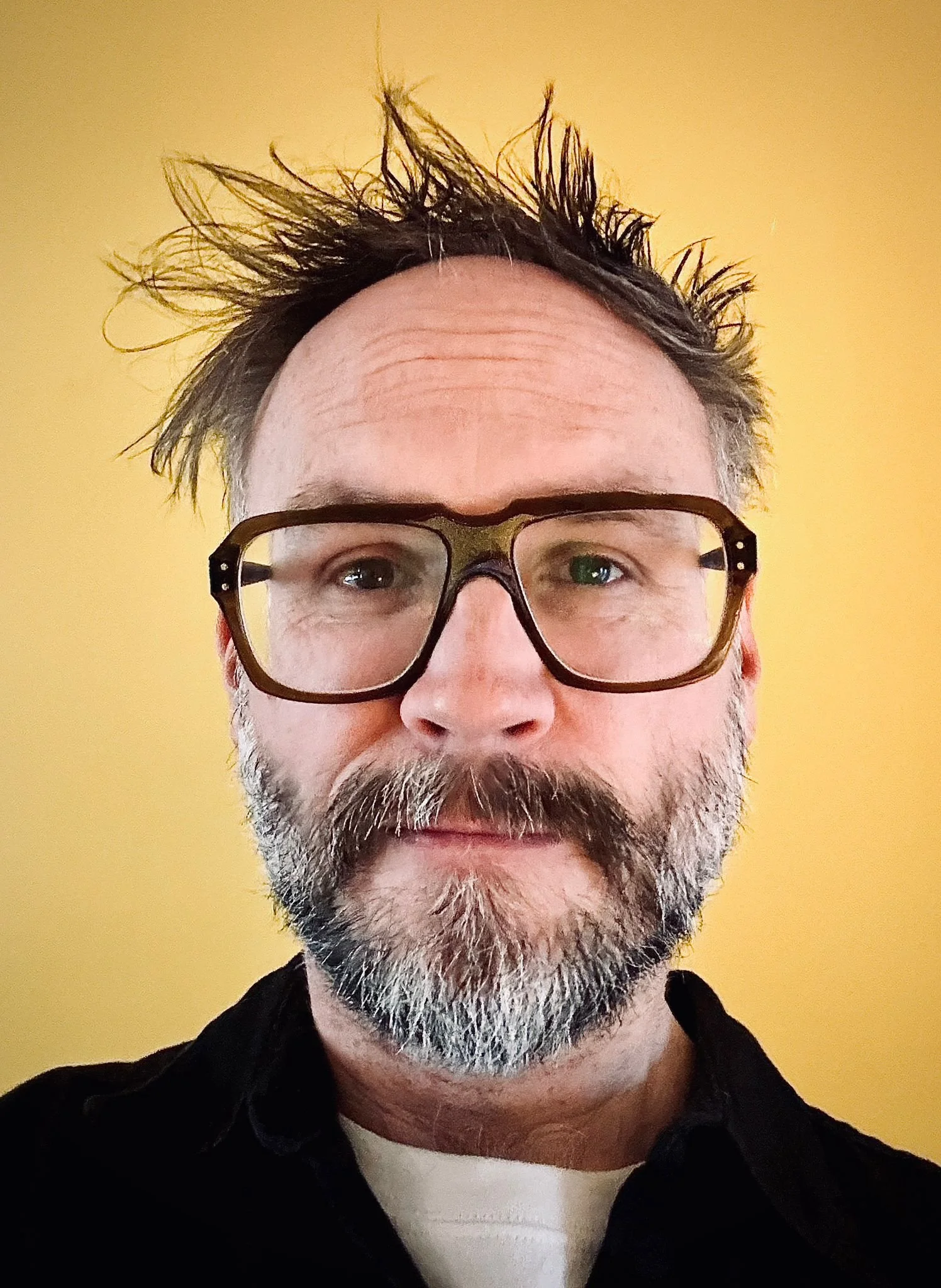 A middle-aged man with spiked dark hair, glasses, and a beard, wearing a black hoodie over a white shirt, standing against a yellow background.