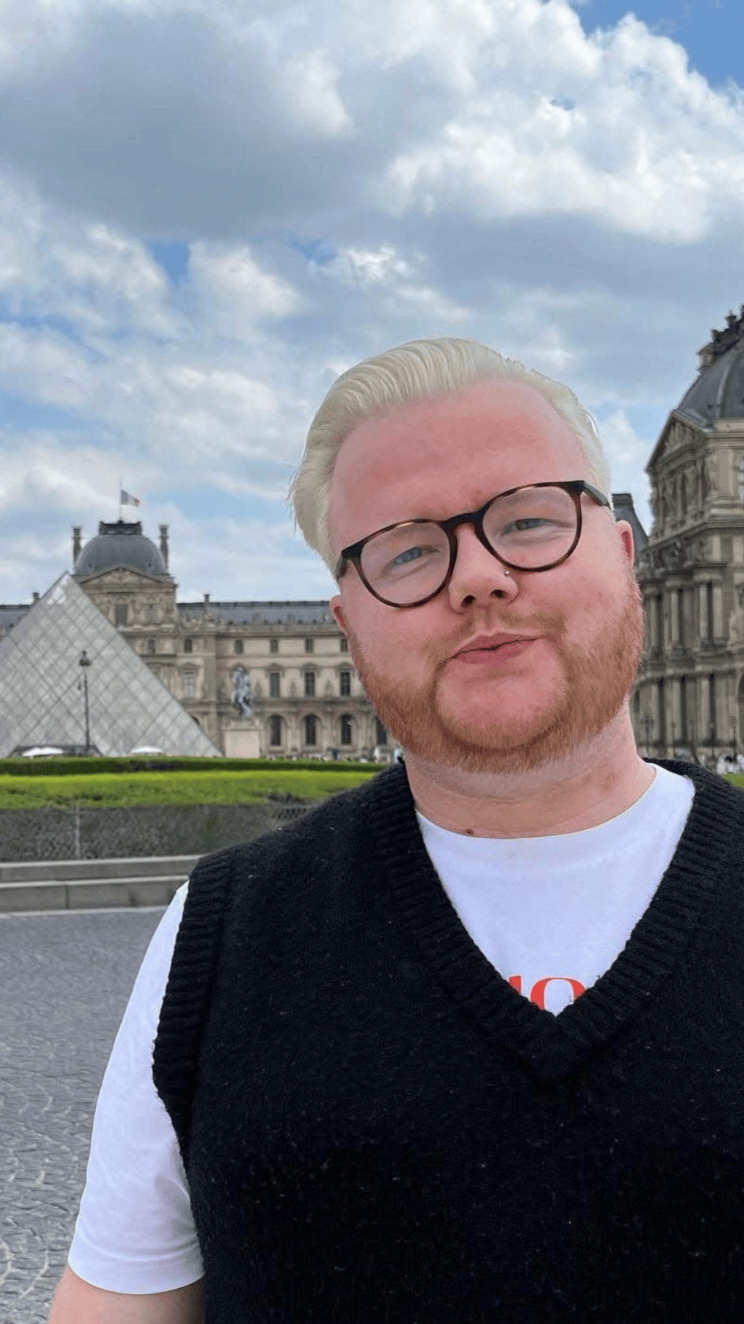 A person with blonde hair, glasses, and a red beard taking a selfie in front of the Louvre Museum in Paris, France.