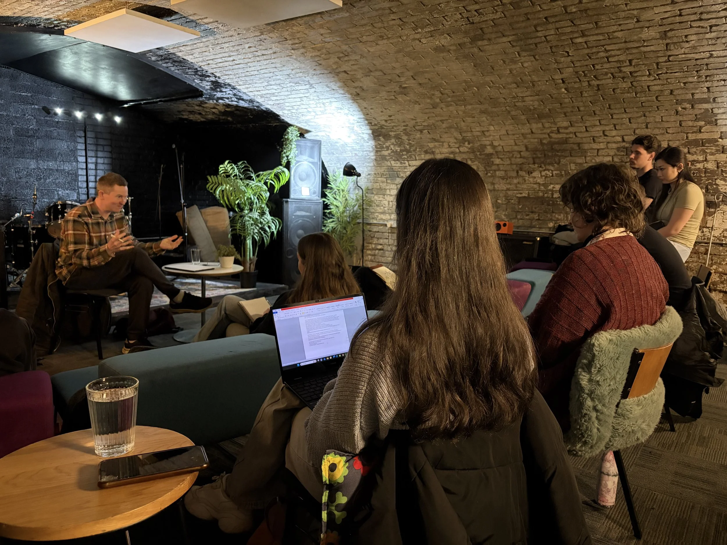 A group of people gathered in a dimly lit room with exposed brick walls, sitting and listening to a speaker. The speaker, a man in a plaid shirt, is seated on a chair, gesturing with his hands. There are musical instruments, plants, and stage equipment in the background, indicating a casual event or discussion.