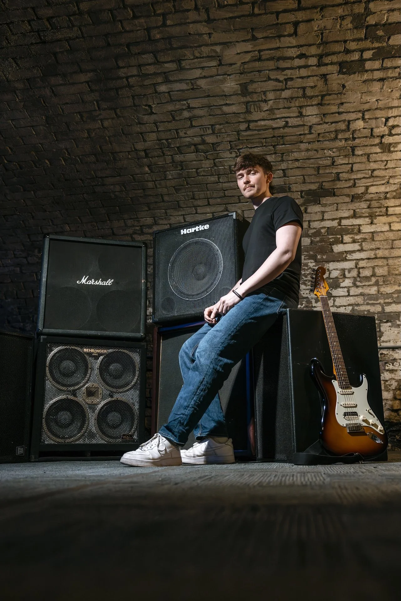 A young man in a black t-shirt and jeans sitting on a stack of guitar amplifiers in a room with an exposed brick ceiling. To his right, an electric guitar is leaning against one of the amplifiers.