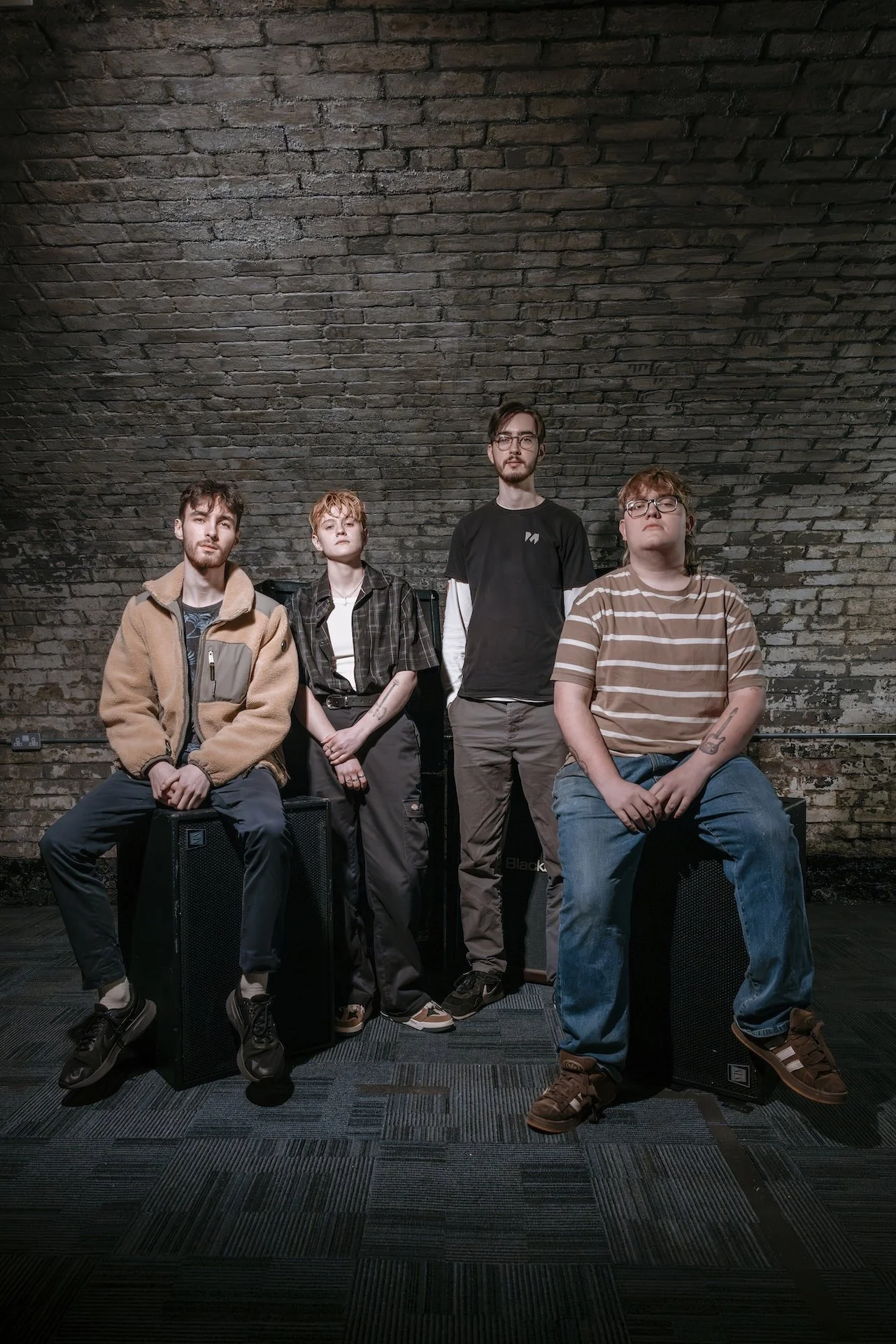 Sugar Bang Press Photo - Four young men with varying hairstyles and glasses pose against a brick wall in a studio setting, with two sitting and two standing, all dressed casually.