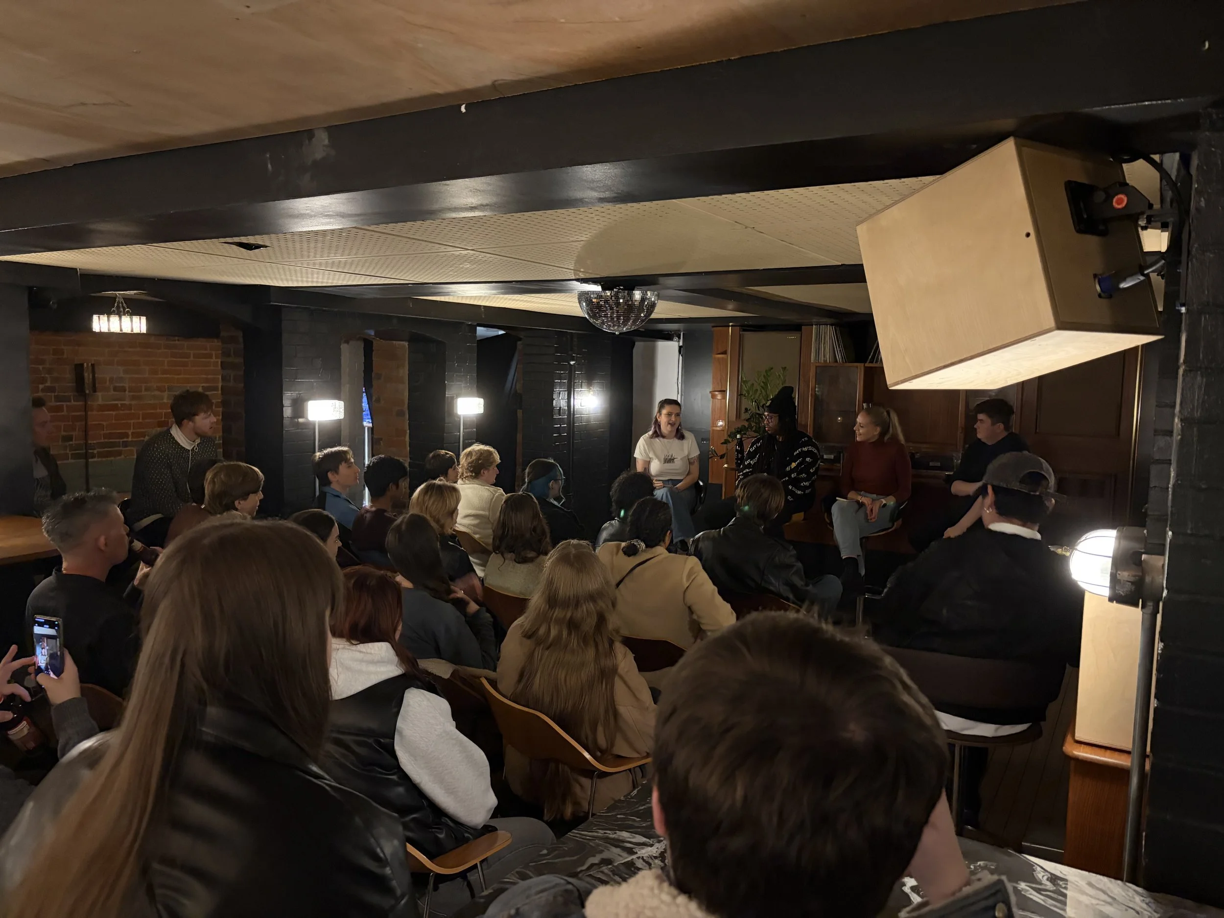 A group of people gathered in a dimly lit room, sitting and standing, listening to two women speaking in front of the audience. The room has brick walls, wooden furniture, and a disco ball on the ceiling.