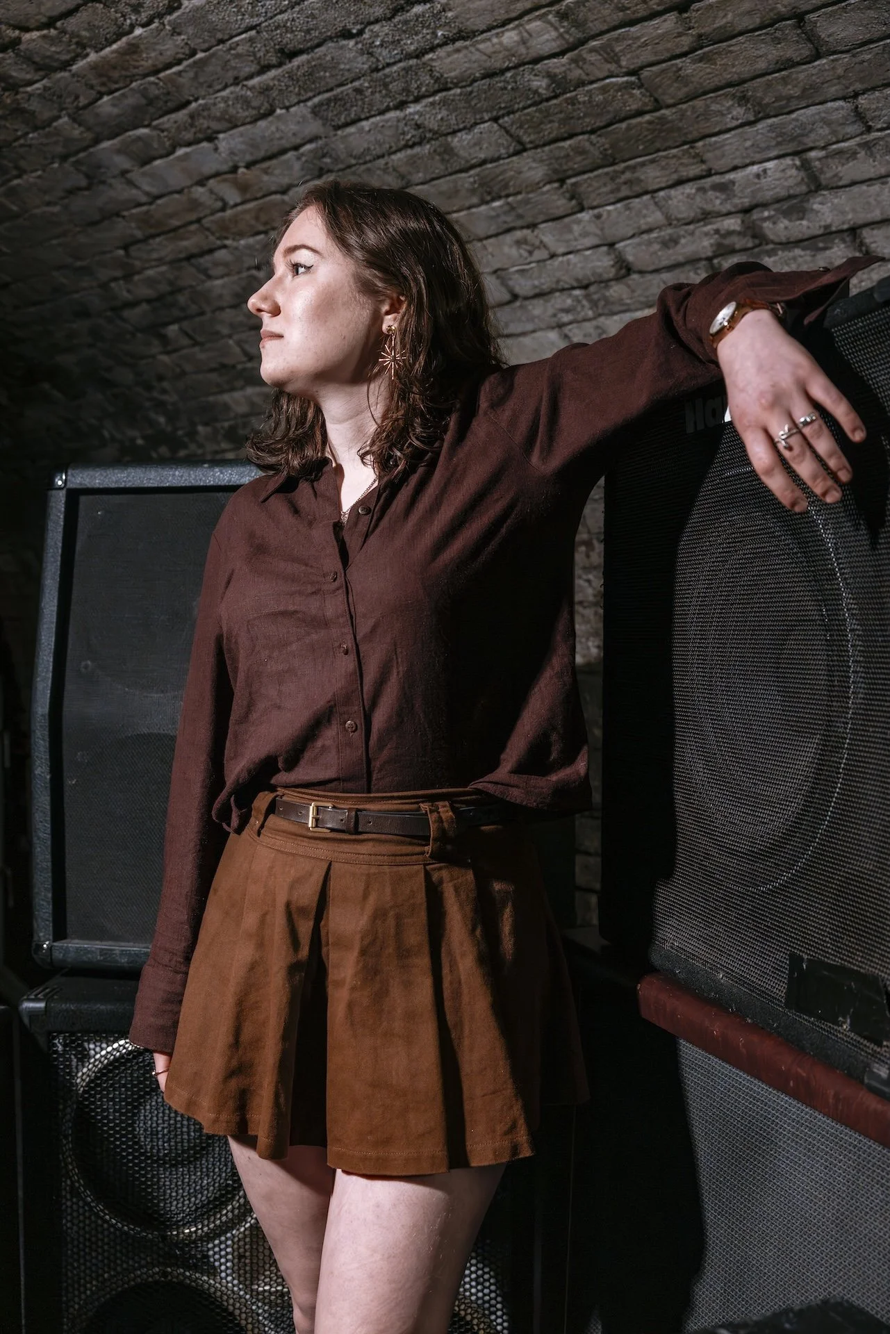 A woman with shoulder-length brown hair wearing a brown shirt and brown pleated skirt, looking to her left, standing in front of a brick wall and surrounded by large black speaker cabinets.