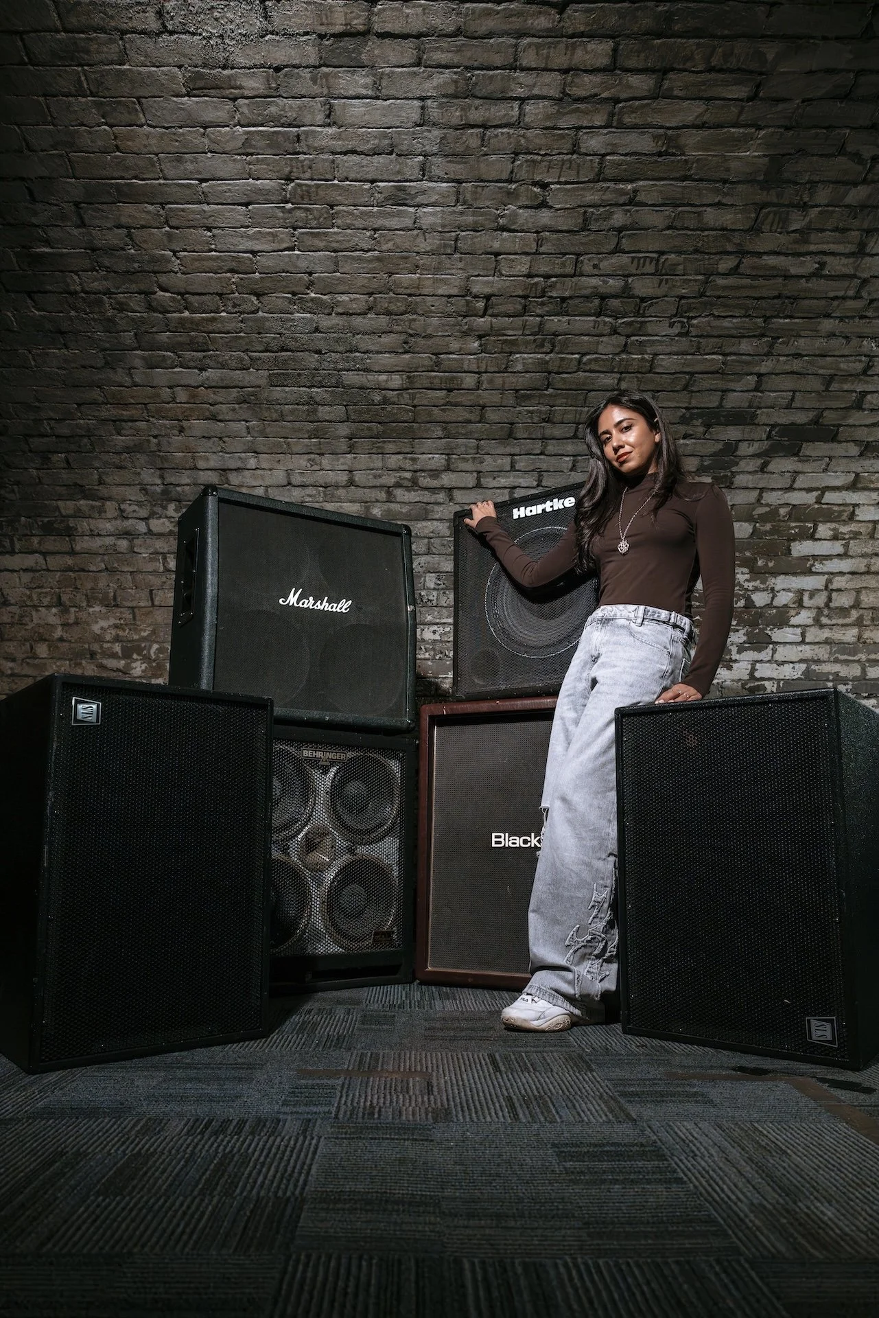 A woman with dark hair wearing a brown long-sleeve shirt, light-colored baggy jeans, and white sneakers, standing among large black speakers with a brick wall background.