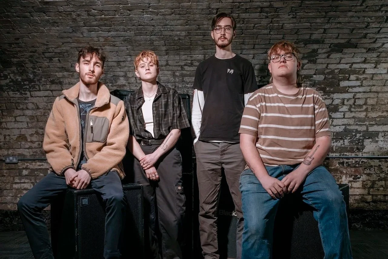 Four young adults sitting and standing against a brick wall, with musical equipment visible.