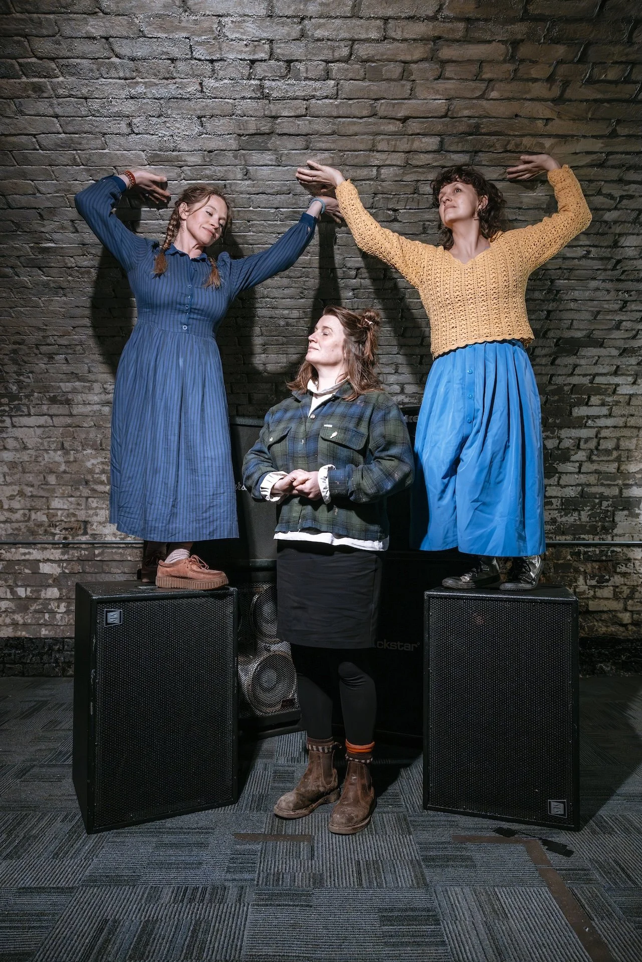 Three women with different hair styles and clothing standing on speakers against a brick wall, engaging in a symbolic pose of unity or strength.