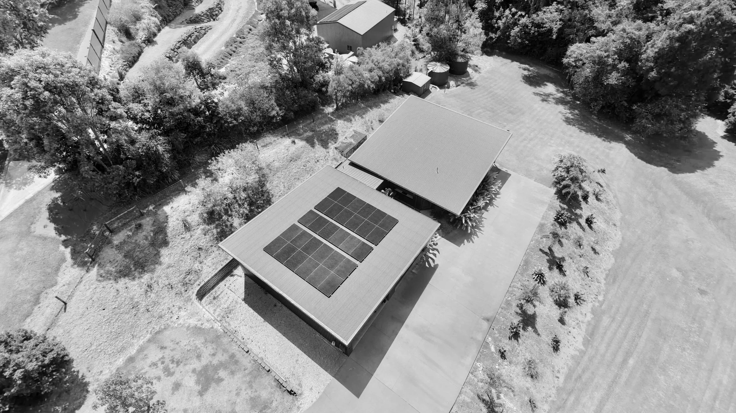 An aerial view of a house with solar panels on the roof, surrounded by trees and a driveway with several parked vehicles.