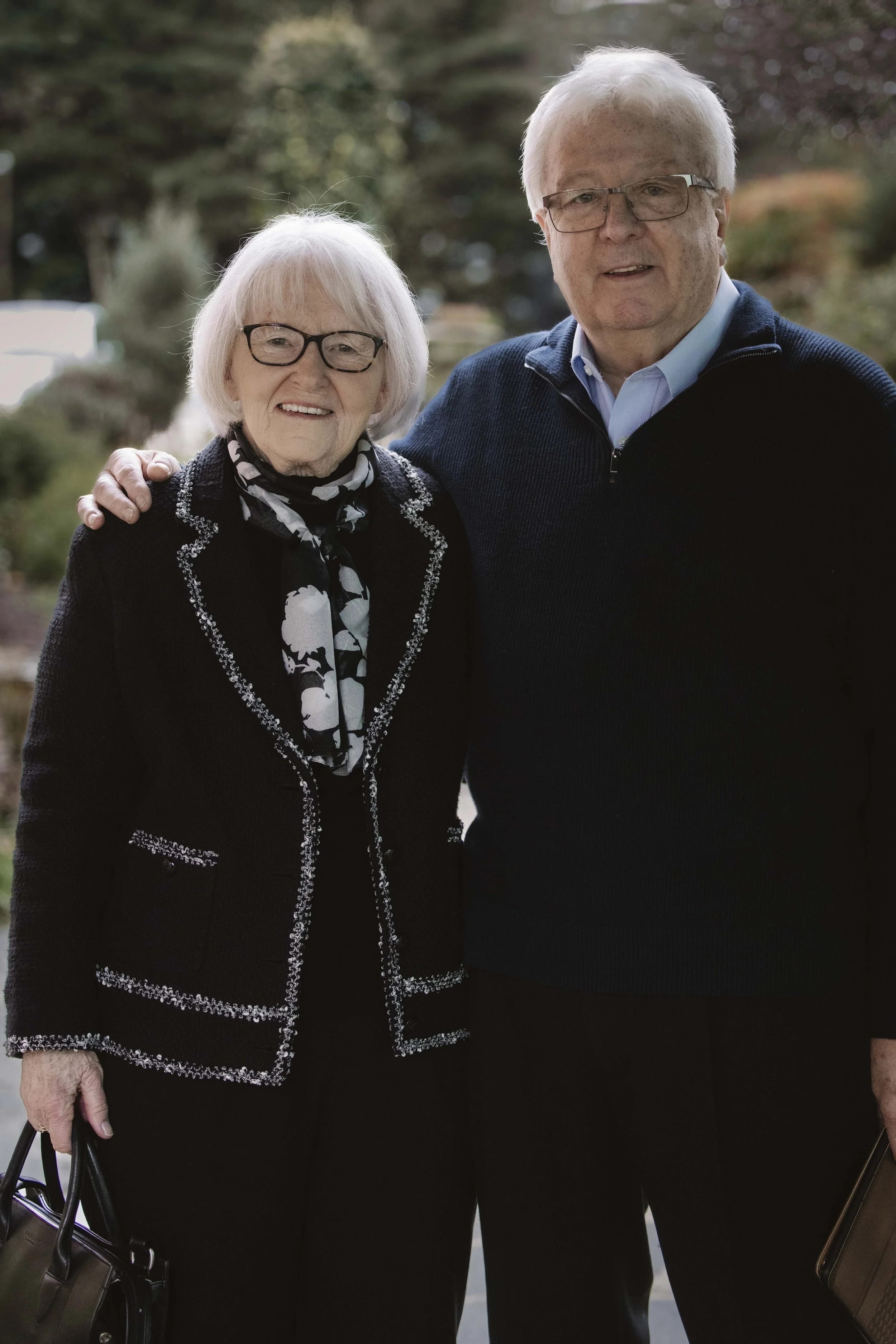 An elderly woman and man standing outdoors together, smiling, with trees in the background.