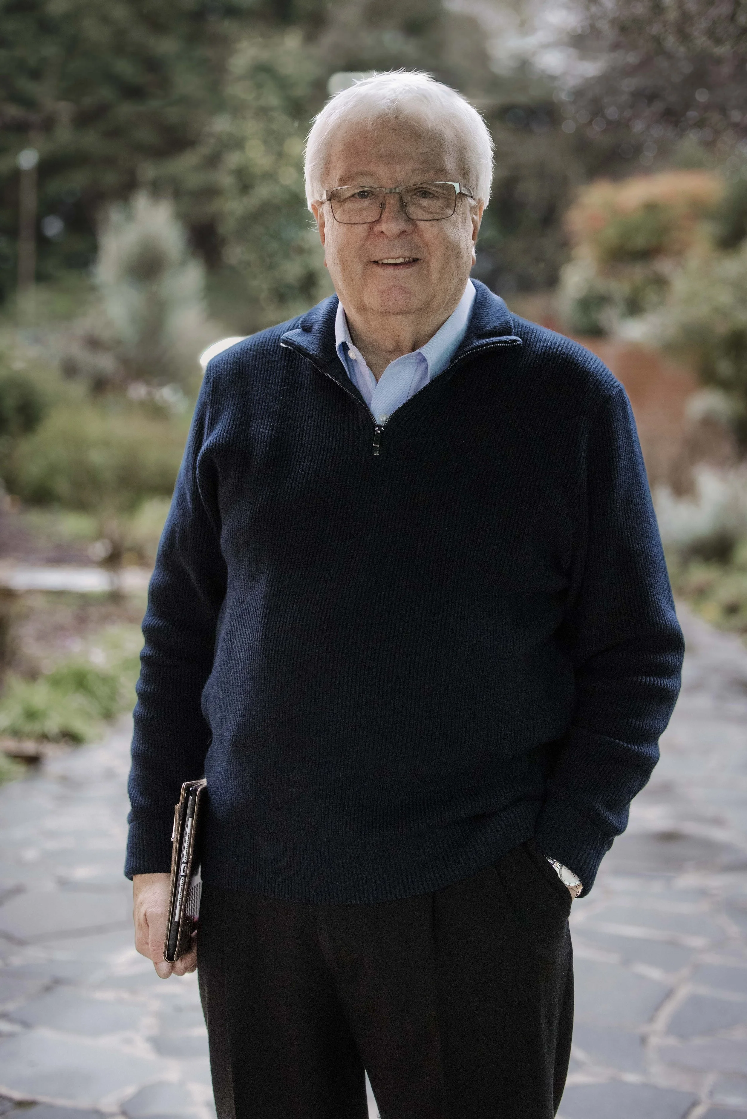 An elderly man with white hair and glasses standing outdoors on a stone pathway with trees and bushes in the background, wearing a navy blue sweater and light blue collared shirt, holding a notebook in his left hand.
