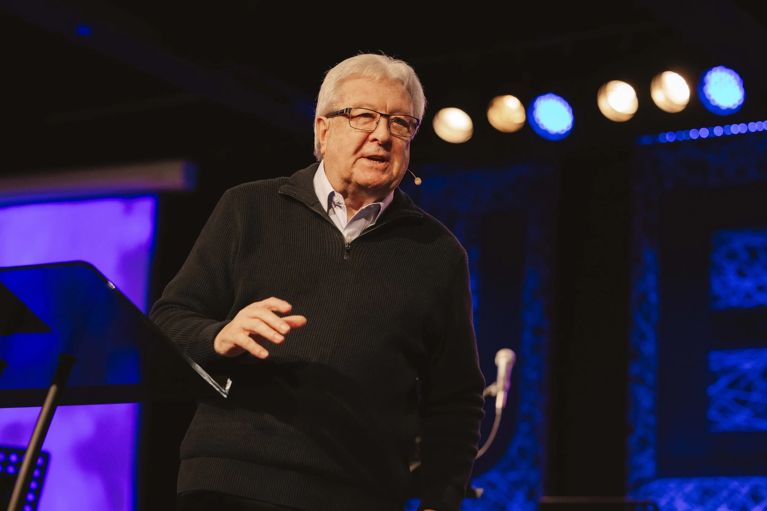 An elderly man with glasses and white hair speaking on stage, with blue and orange stage lights in the background.